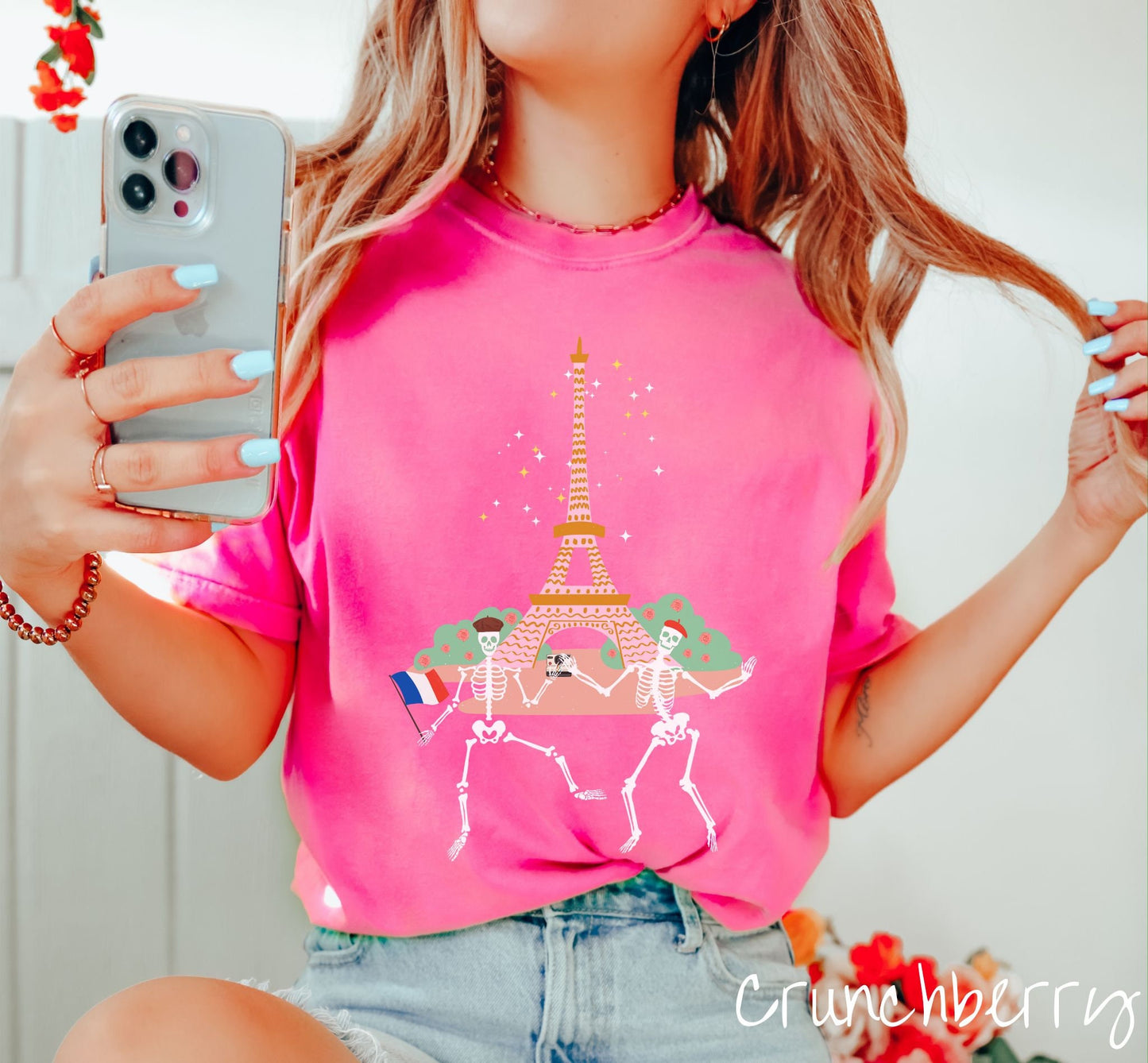 A woman wearing a vintage, blossom colored shirt with two skeletons wearing colorful French berets taking a selfie picture in front of the Eiffel Tower at night in Paris, France with twinkling stars in the background of the tower.