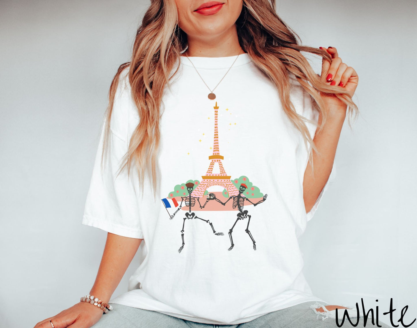 A woman wearing a vintage, white colored shirt with two skeletons wearing colorful French berets taking a selfie picture in front of the Eiffel Tower at night in Paris, France with twinkling stars in the background of the tower.
