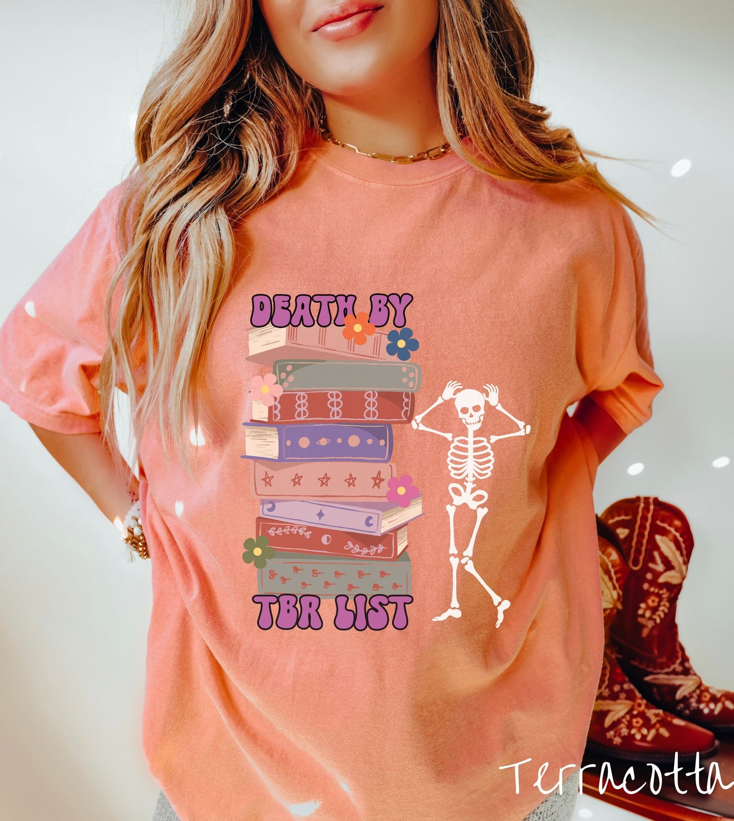 A woman wearing a cute terracotta colored shirt with a huge stack of books with flowers sandwiched by the text Death by TBR List and a skeleton standing next to the stack with its hands on its head.