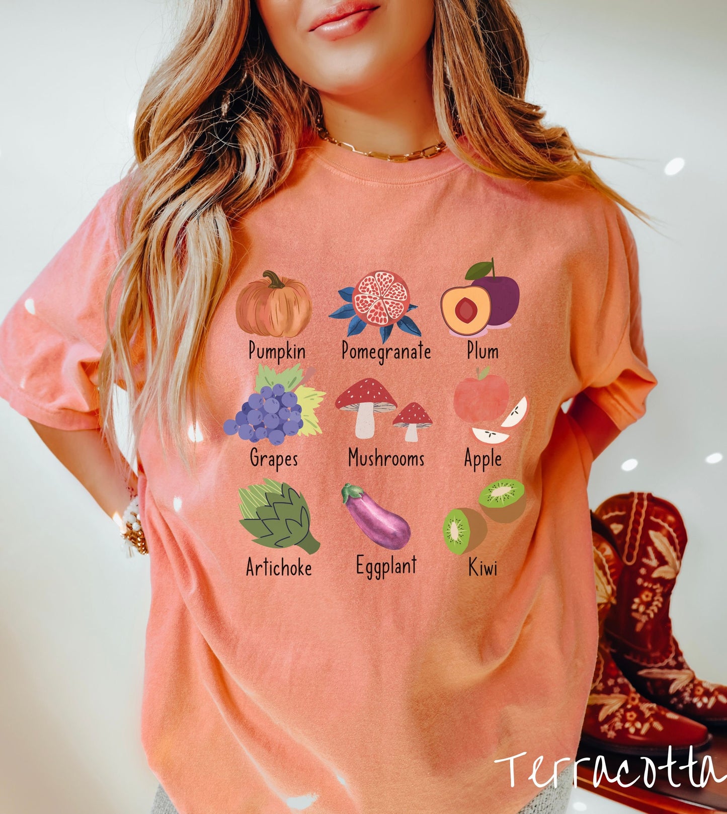 A woman wearing a vintage, terracotta colored shirt with the picture and text of a pumpkin, a pomegranate, a plum, grapes, mushrooms, an apple, an artichoke, an eggplant, and a kiwi listed in three rows of three.