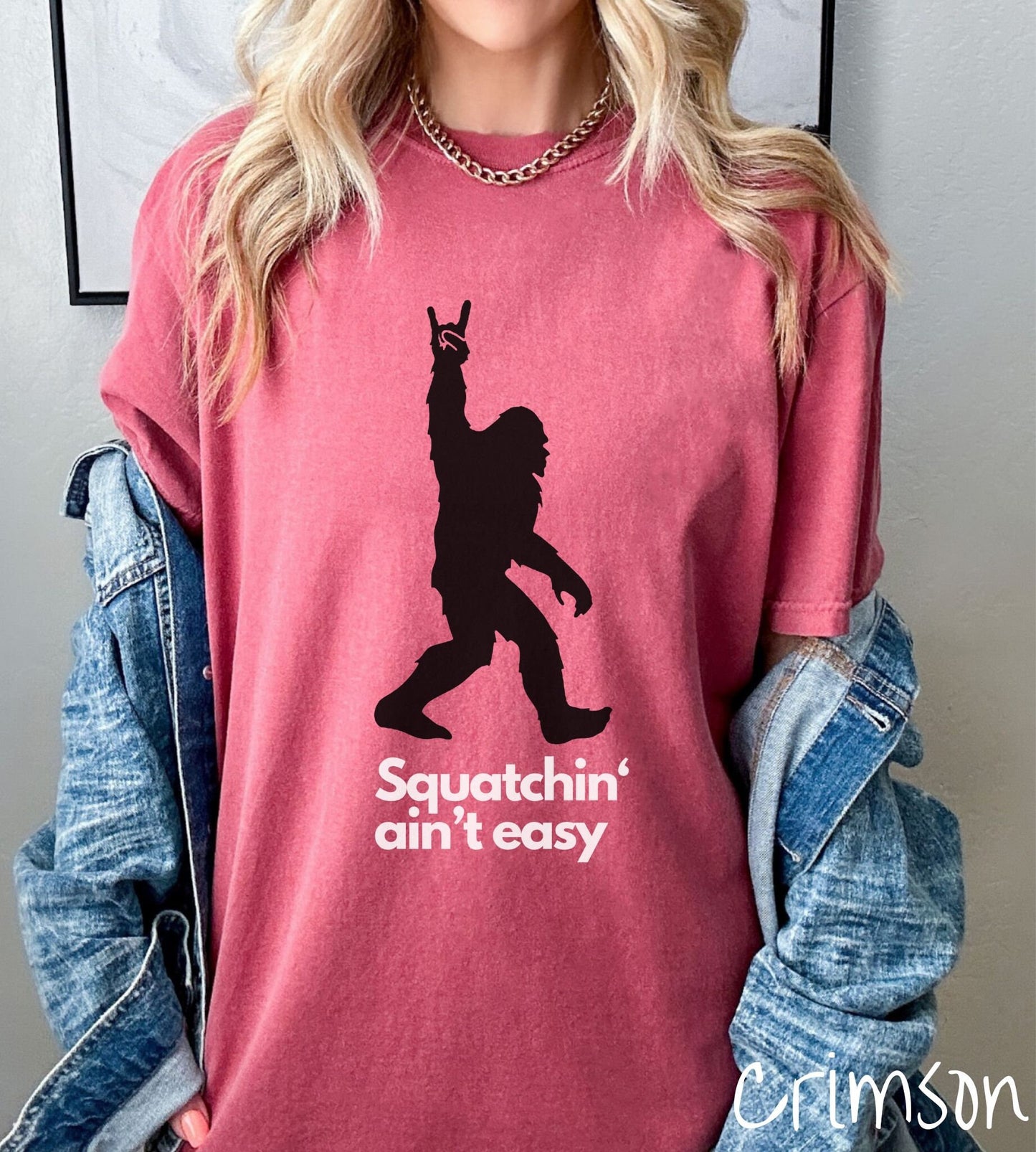 A woman wearing a cute crimson colored shirt with text saying Squatchin aint easy and a silhouette of Bigfoot holding up the rock on hand sign.
