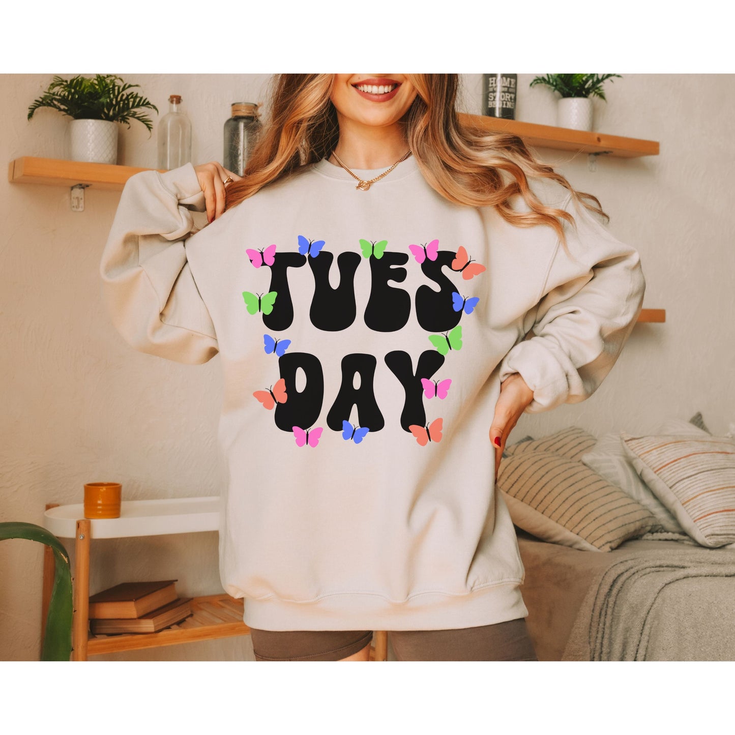 A woman wearing a vintage, sand colored sweatshirt with the text centered on the front TUES then DAY below it, with pink, green, blue, and orange butterflies scattered amongst the letters.