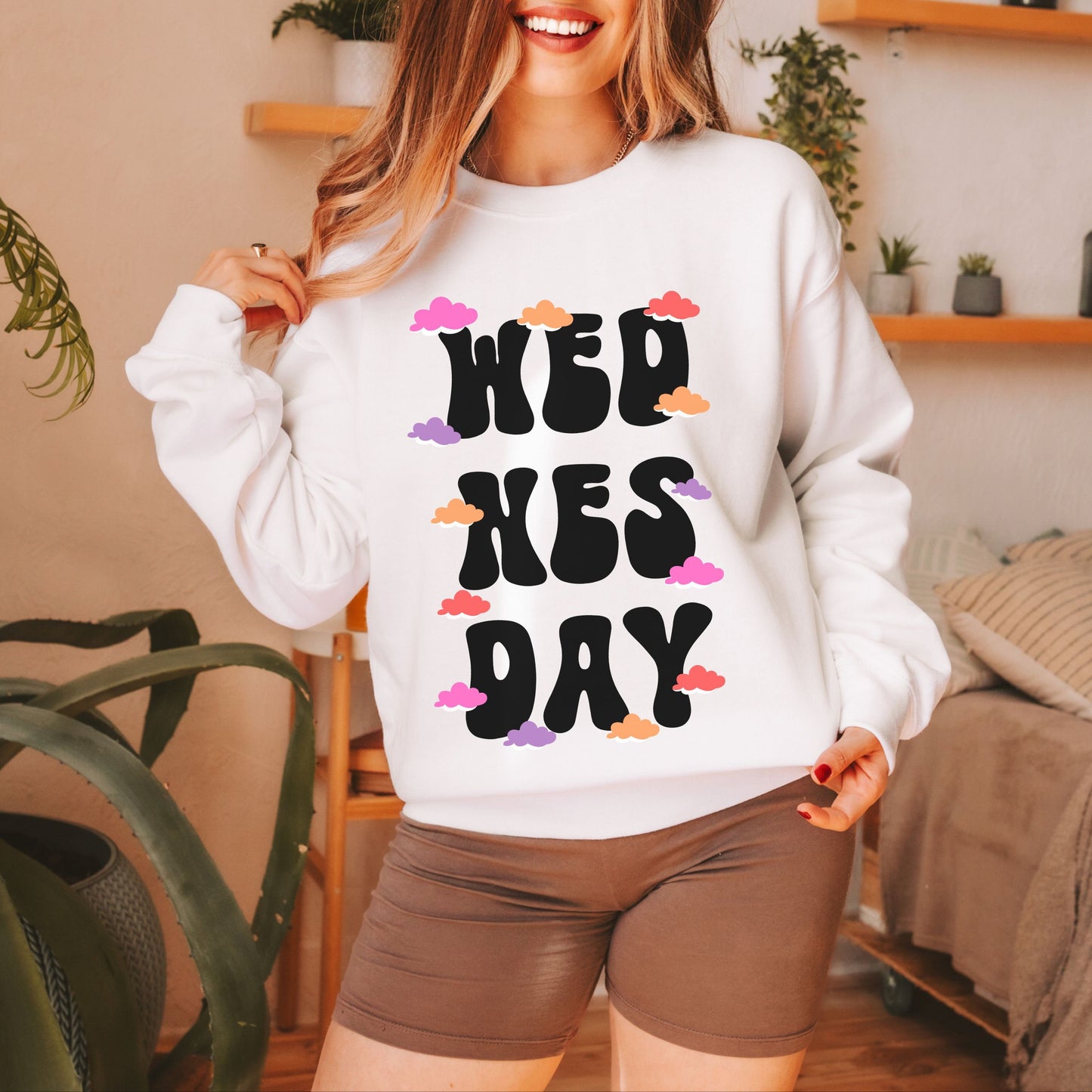 A woman wearing a vintage, white colored sweatshirt with the text centered on the front WED then below it NES then DAY below that, with pink, red, purple, and orange clouds scattered amongst the letters.