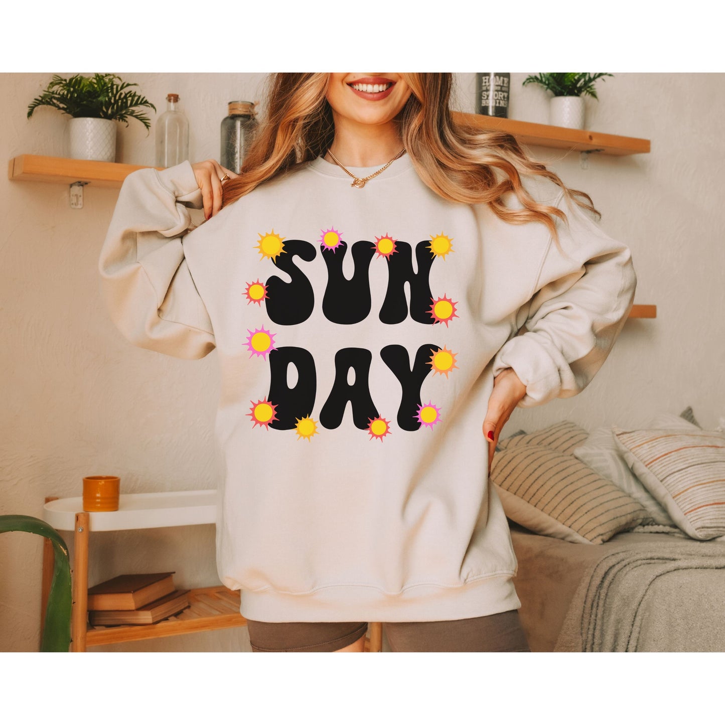 A woman wearing a vintage, sand colored sweatshirt with the text centered on the front SUN then below it DAY, with yellow, orange, and pink suns scattered amongst the letters.