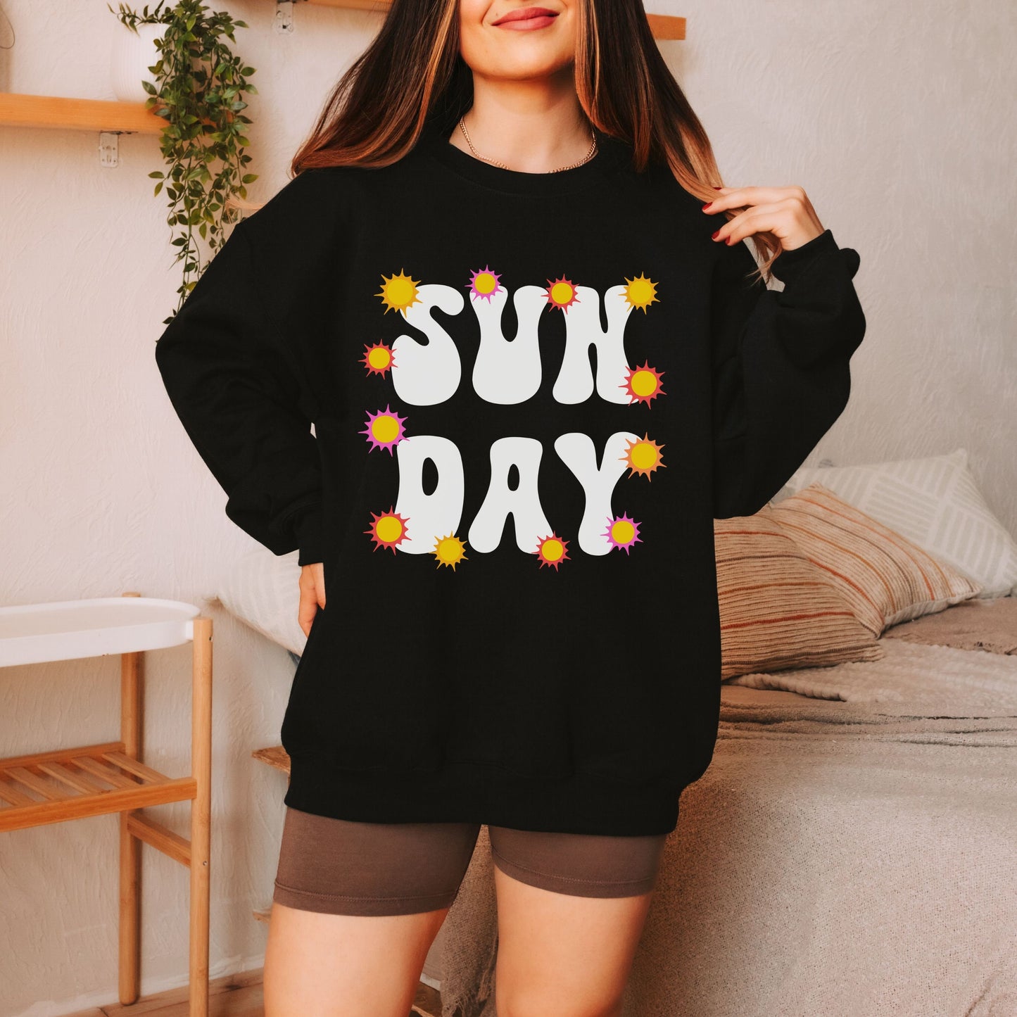A woman wearing a vintage, black colored sweatshirt with the text centered on the front SUN then below it DAY, with yellow, orange, and pink suns scattered amongst the letters.