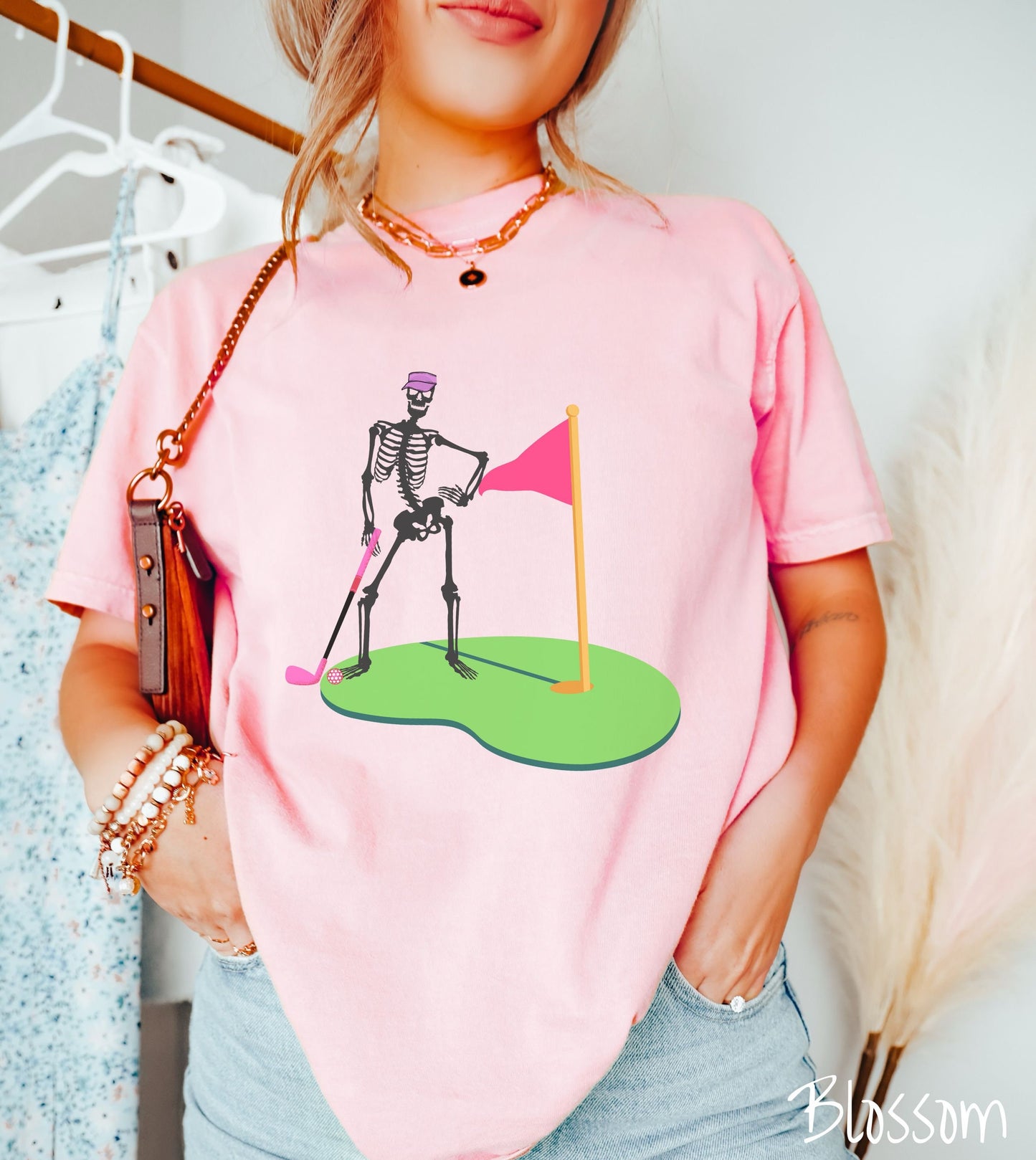 A woman wearing a cute blossom colored shirt with a black skeleton posing on the golfing green with a pink putter, pink golf ball, and a pink flag in the hole.