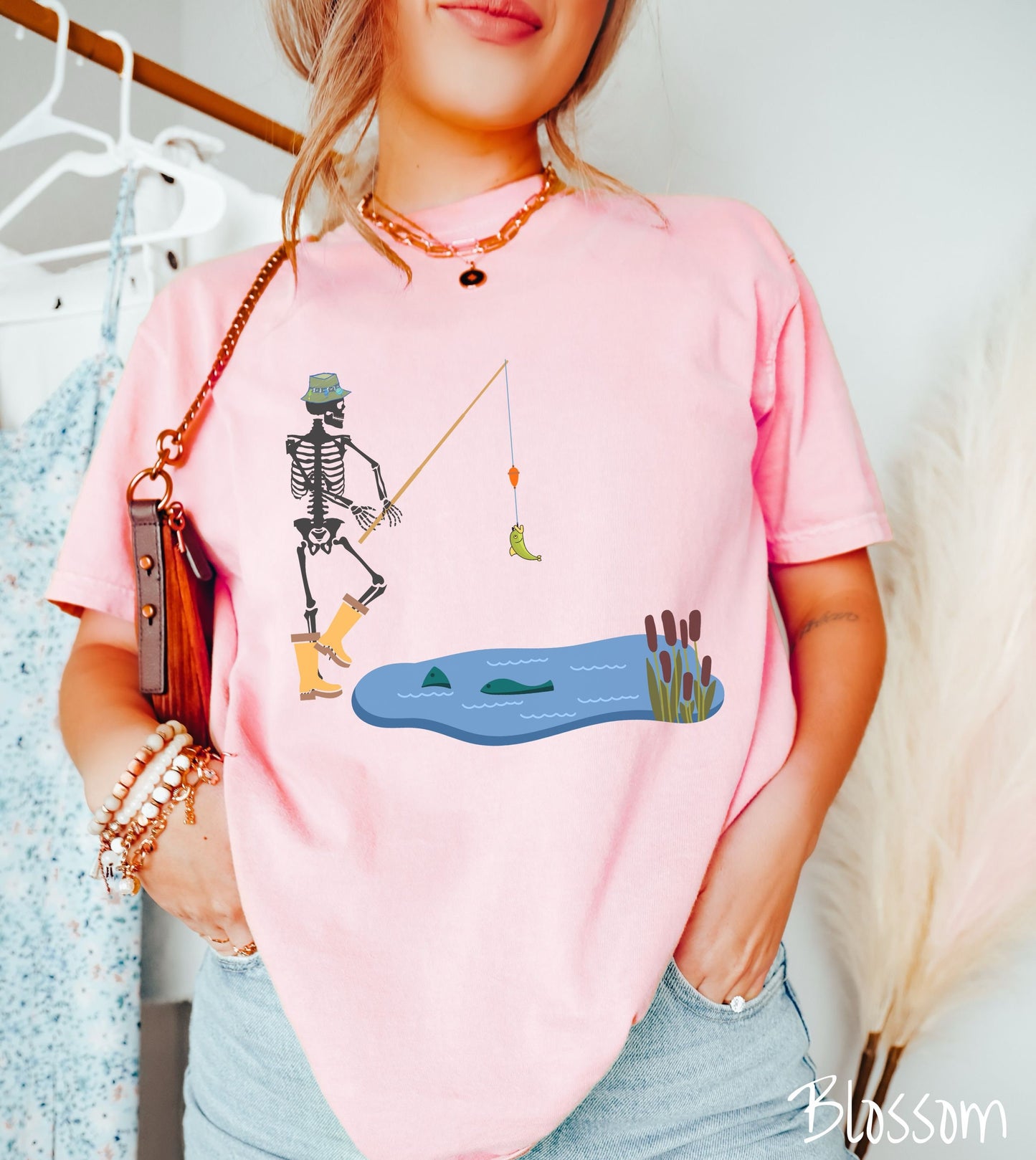 A woman wearing a cute, vintage blossom colored shirt with a skeleton wearing yellow rain boots reeling in a green bass from a pond with fish swimming at the top.