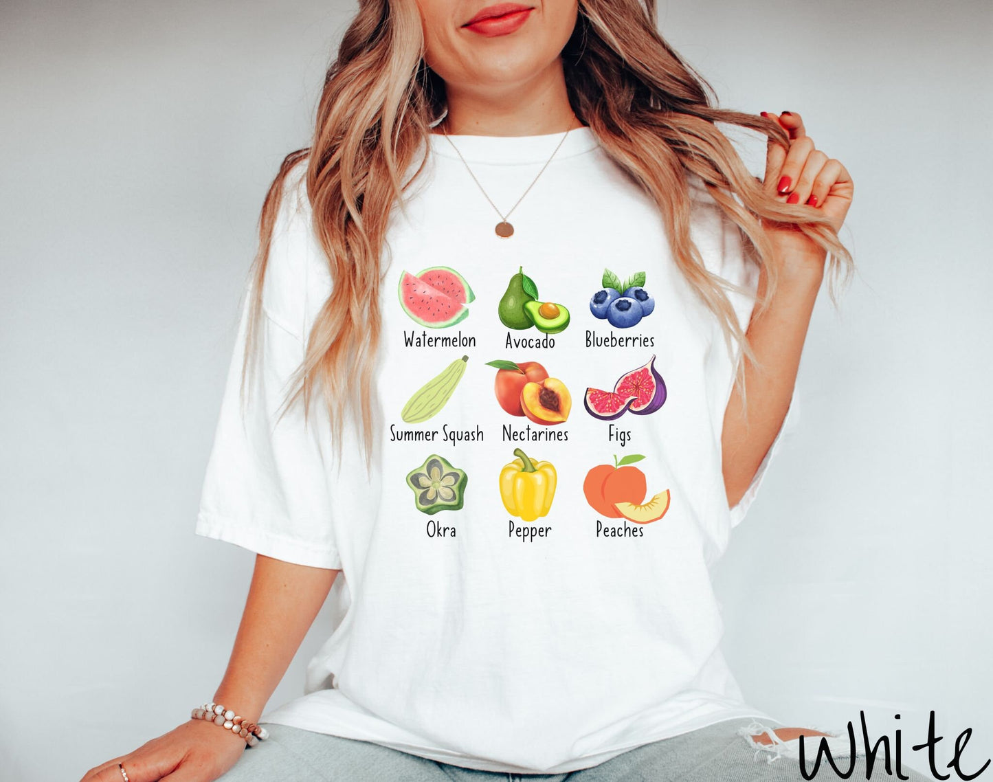 A woman wearing a vintage, white colored shirt with the picture and text of a watermelon, an avocado, blueberries, a summer squash, nectarines, figs, oka, a pepper, and peaches listed in three rows of three.