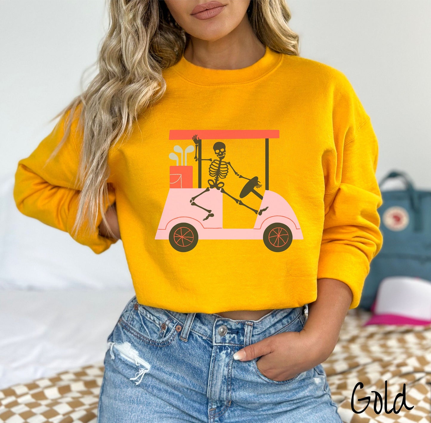 A woman wearing a cute gold colored sweatshirt with a skeleton wearing a Santa hat driving a pink golf cart and waving.