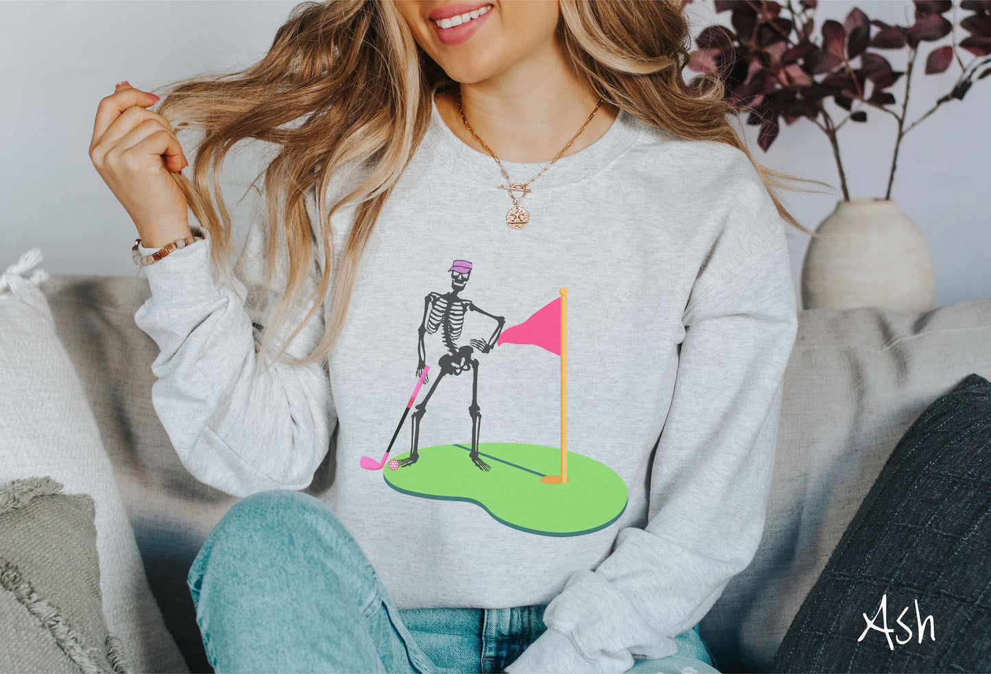 A woman wearing a cute ash colored sweatshirt with a black skeleton posing on the golfing green with a pink putter, pink golf ball, and a pink flag in the hole.