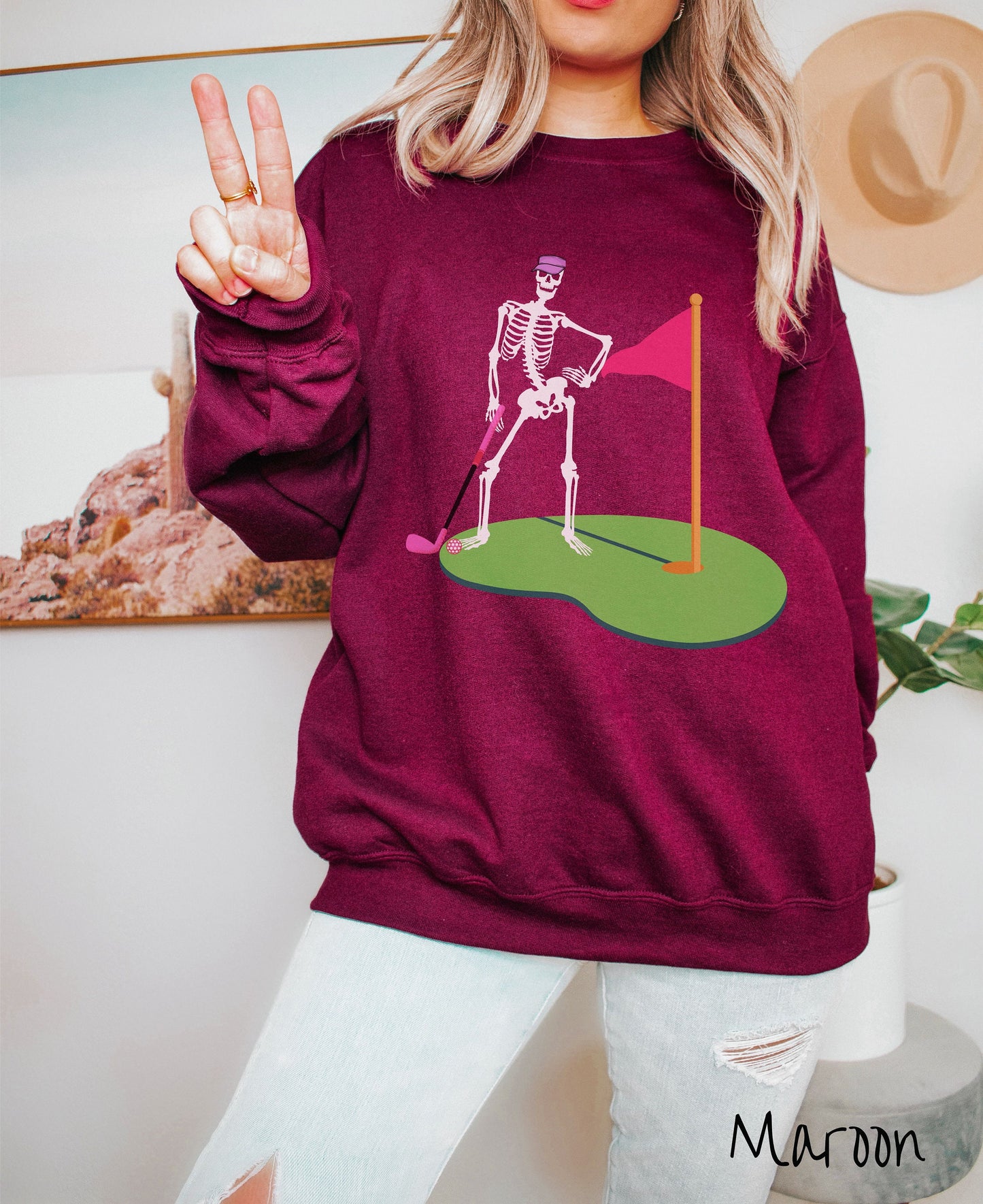 A woman wearing a cute maroon colored sweatshirt with a black skeleton posing on the golfing green with a pink putter, pink golf ball, and a pink flag in the hole.