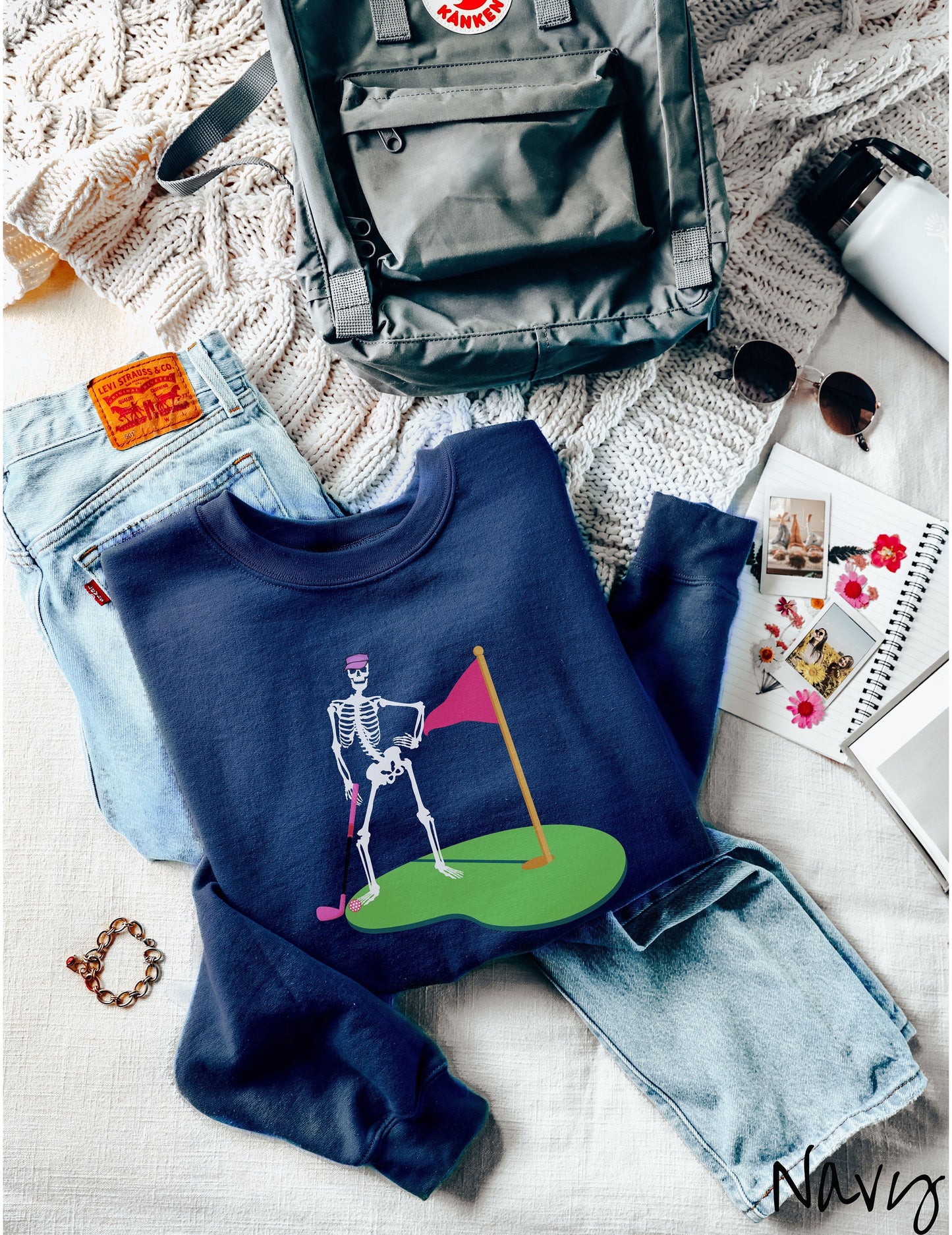 A cute navy colored sweatshirt with a black skeleton posing on the golfing green with a pink putter, pink golf ball, and a pink flag in the hole.