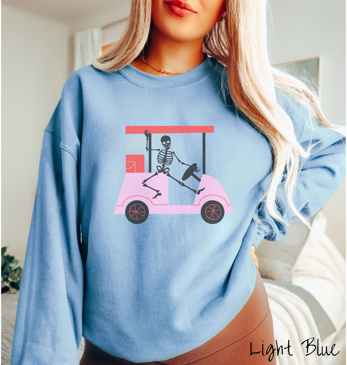 A woman wearing a cute light blue colored sweatshirt with a skeleton wearing a Santa hat driving a pink golf cart and waving.