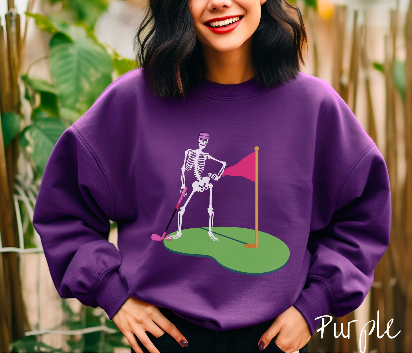 A woman wearing a cute purple colored shirt with a black skeleton posing on the golfing green with a pink putter, pink golf ball, and a pink flag in the hole.