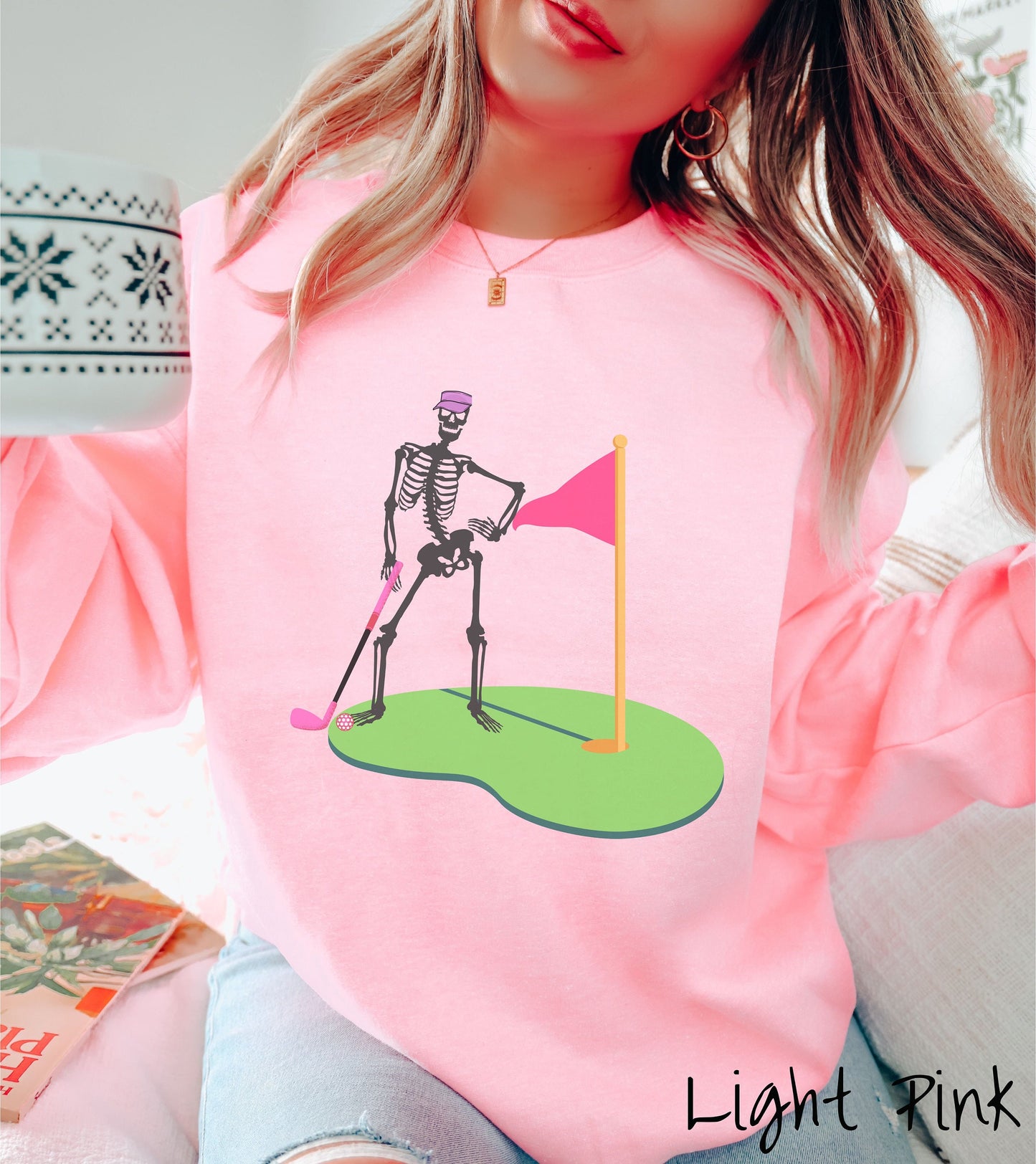 A woman wearing a cute light pink colored sweatshirt with a black skeleton posing on the golfing green with a pink putter, pink golf ball, and a pink flag in the hole.