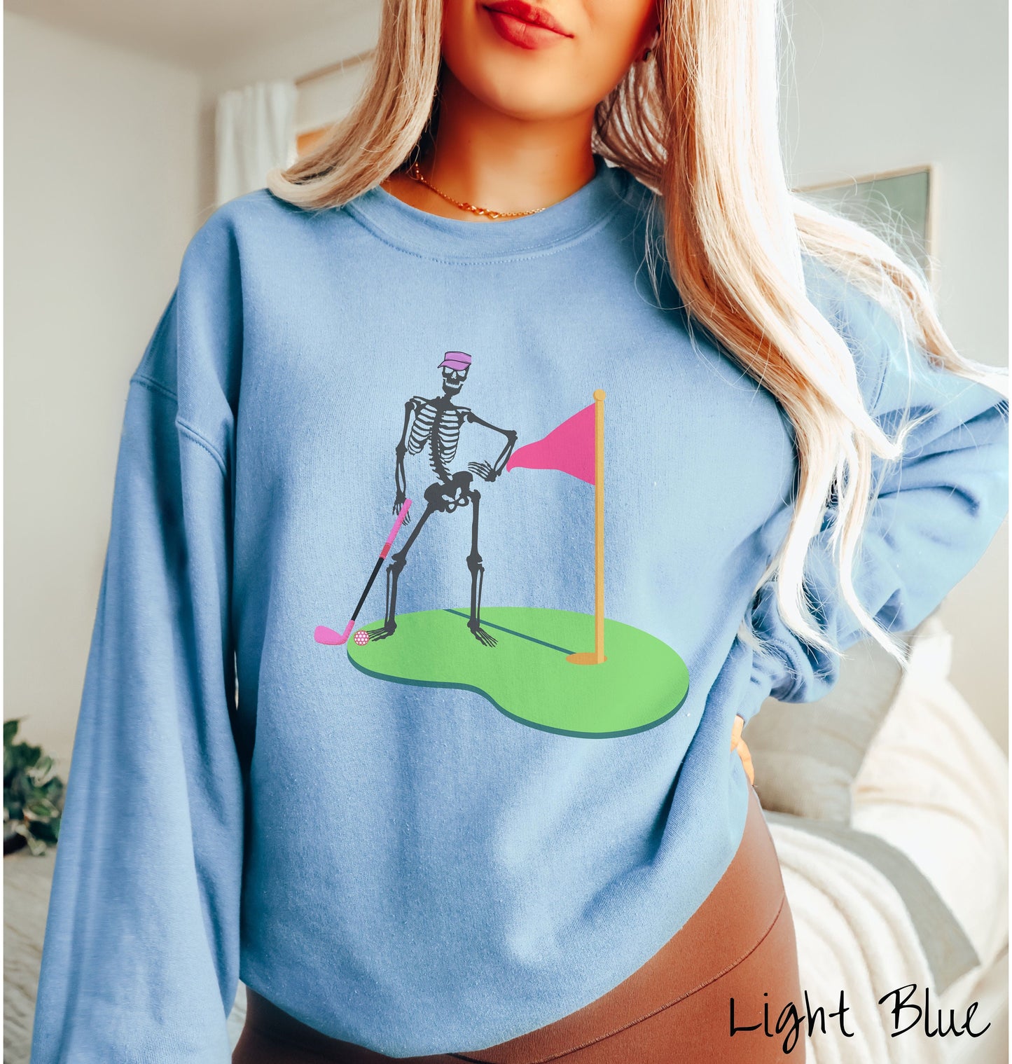A woman wearing a cute light blue colored sweatshirt with a black skeleton posing on the golfing green with a pink putter, pink golf ball, and a pink flag in the hole.