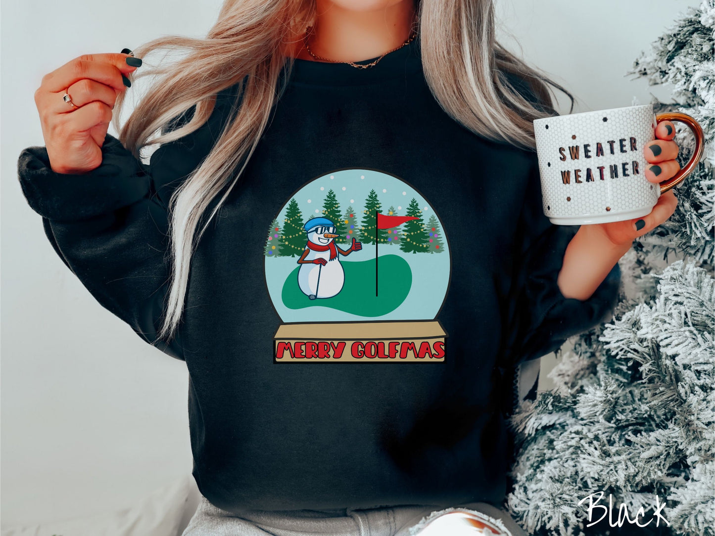 A woman wearing a cute black colored sweatshirt with red text saying Merry Golfmas and a picture of a snowman giving the thumbs up on the golf green next to the hole with a flag in it.