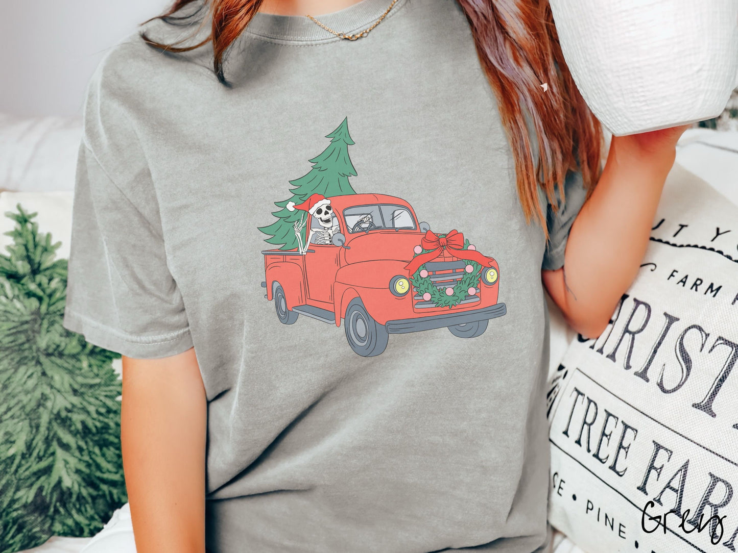 A woman wearing a cute, vintage grey colored comfy sweatshirt with a skeleton wearing a Christmas santa hat driving an old red pickup truck with a Christmas tree stuffed into the back, and a wreath across the front bumper.