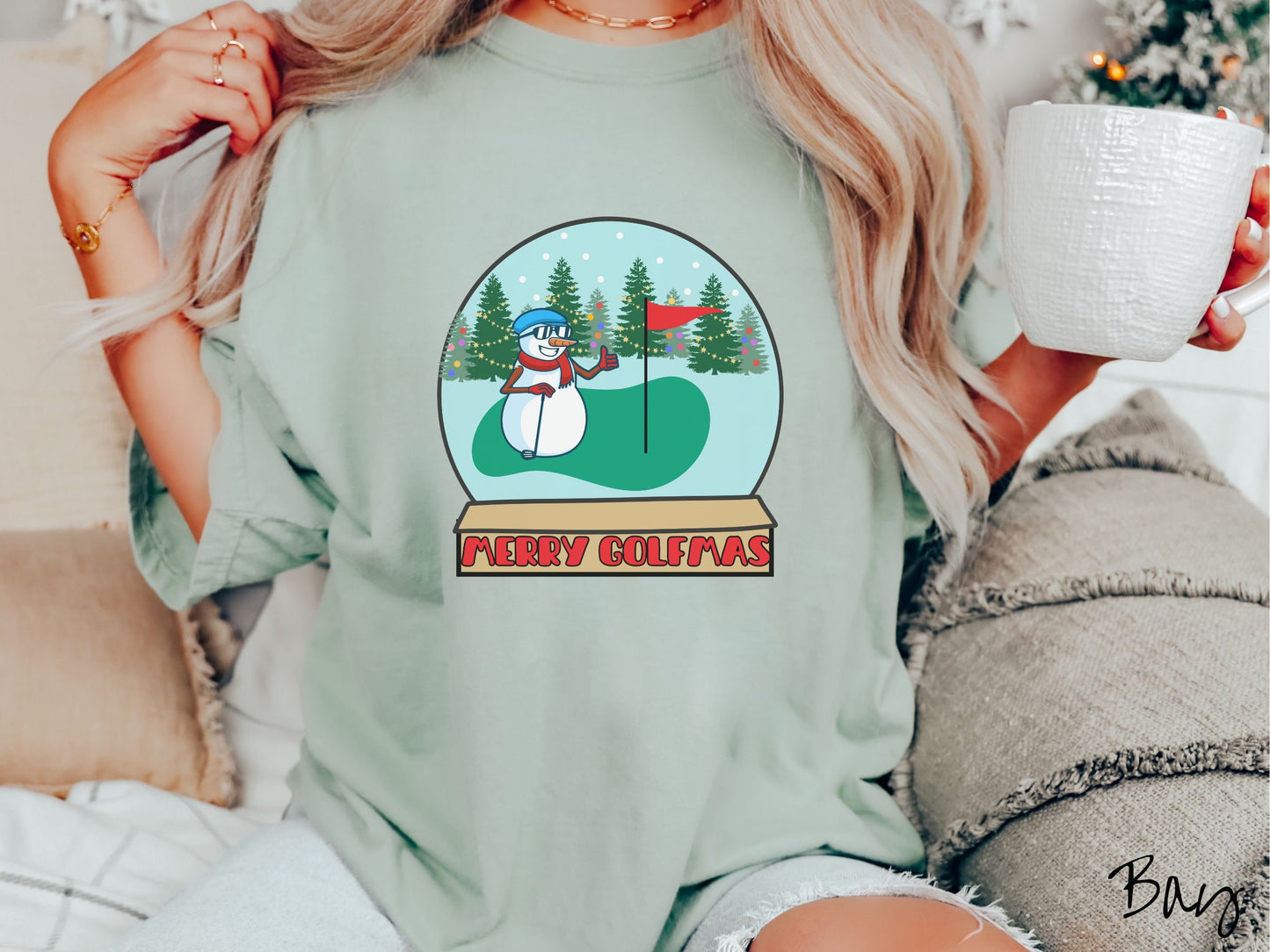 A woman wearing a cute bay colored shirt with red text saying Merry Golfmas and a picture of a snowman giving the thumbs up on the golf green next to the hole with a flag in it.