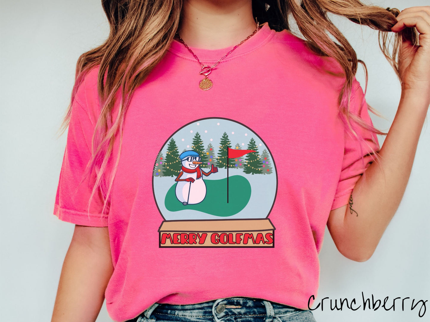 A woman wearing a cute crunchberry colored shirt with red text saying Merry Golfmas and a picture of a snowman giving the thumbs up on the golf green next to the hole with a flag in it.
