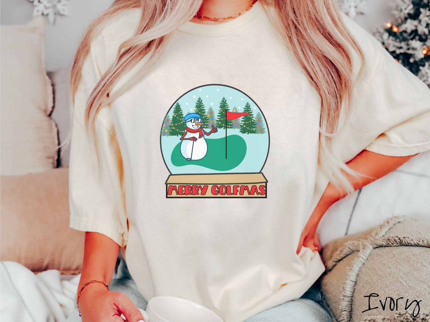 A woman wearing a cute ivory colored shirt with red text saying Merry Golfmas and a picture of a snowman giving the thumbs up on the golf green next to the hole with a flag in it.
