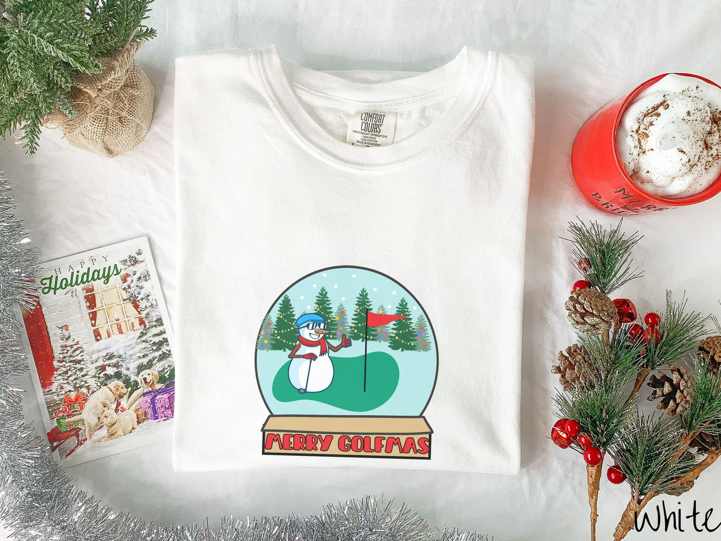A woman wearing a cute white colored shirt with red text saying Merry Golfmas and a picture of a snowman giving the thumbs up on the golf green next to the hole with a flag in it.