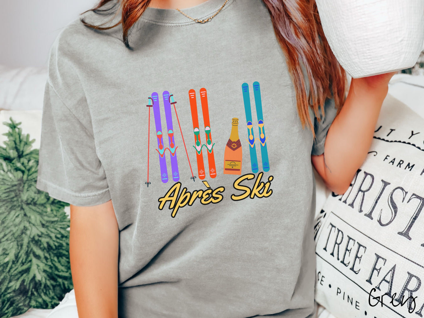 A woman wearing a vintage, grey colored shirt with sets of purple, orange, and turquoise snow skis standing upright, and a big bottle of champagne next to them. Below the skis is the text Apres Ski which is French for After Skiing.