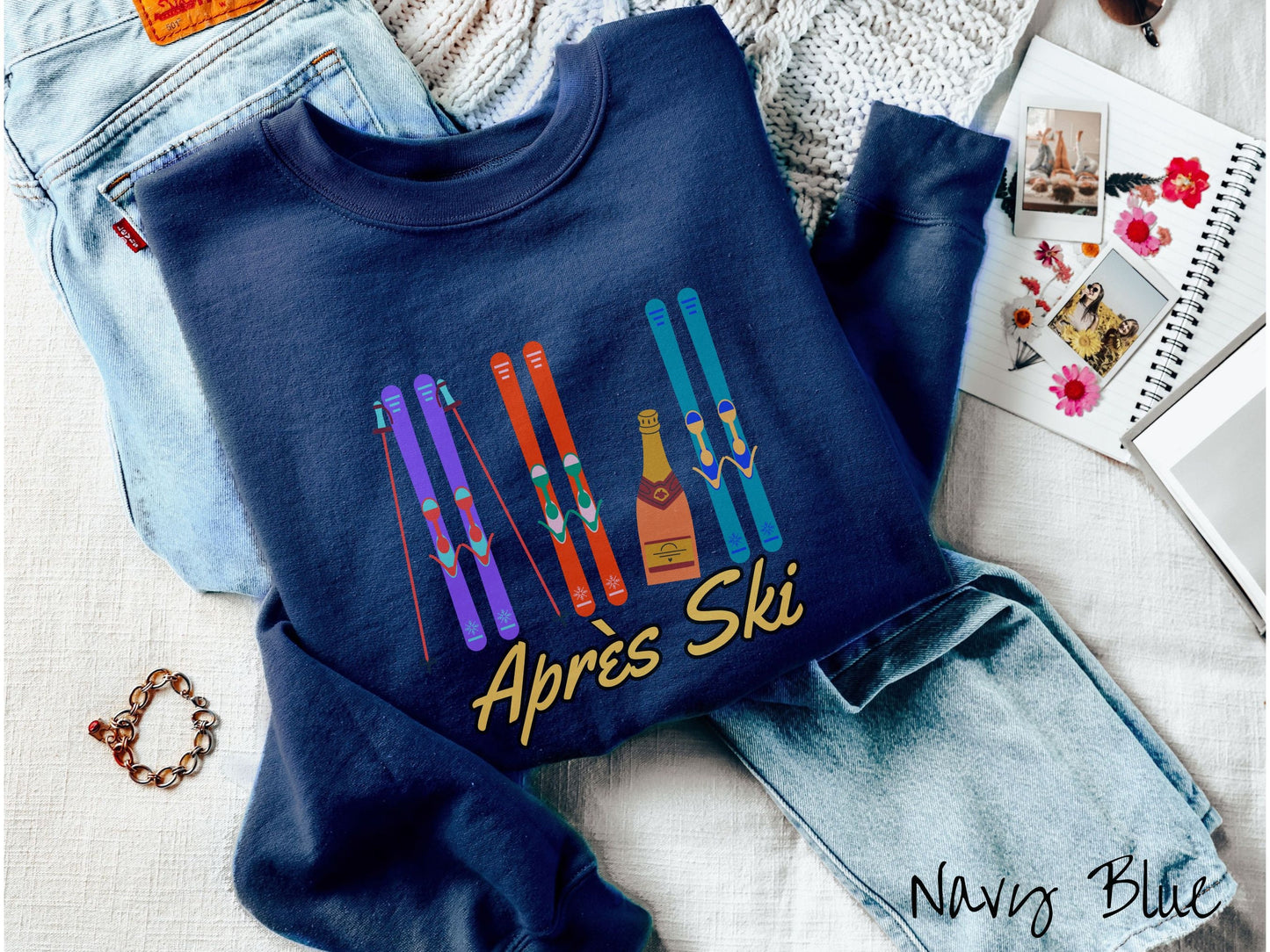 A woman wearing a vintage, navy blue colored sweatshirt with sets of purple, orange, and turquoise snow skis standing upright, and a big bottle of champagne next to them. Below the skis is the text Apres Ski which is French for After Skiing.