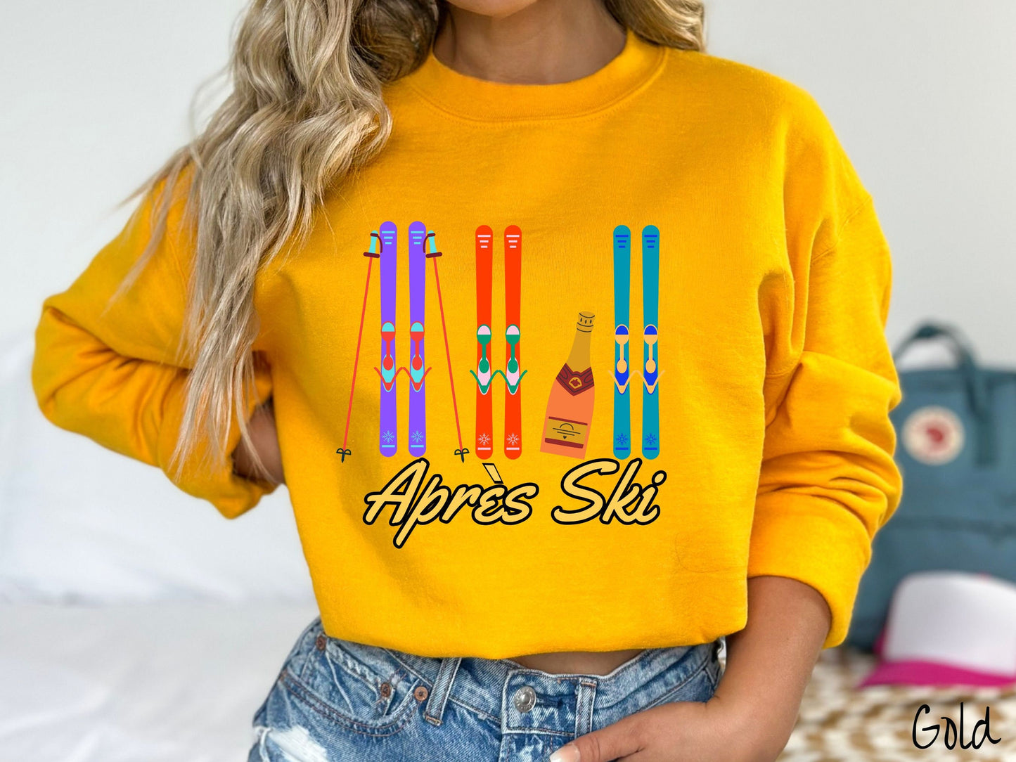 A woman wearing a vintage, gold colored sweatshirt with sets of purple, orange, and turquoise snow skis standing upright, and a big bottle of champagne next to them. Below the skis is the text Apres Ski which is French for After Skiing.
