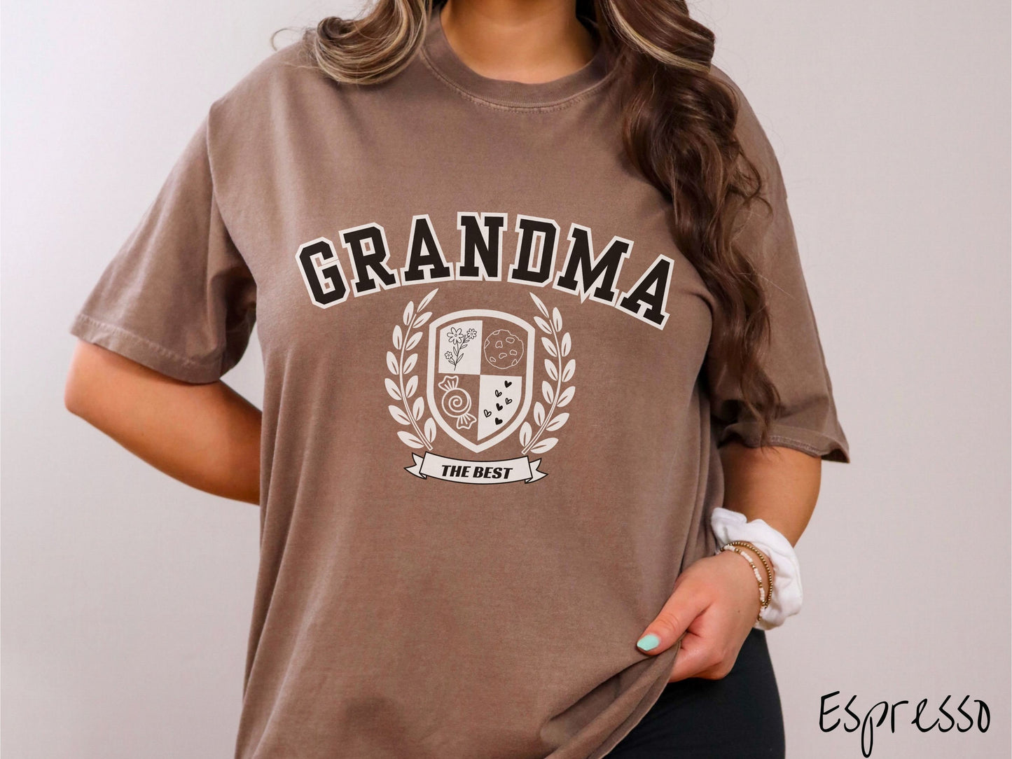 A woman wearing a cute espresso colored shirt with text saying Grandma across the front with a varsity logo underneath with the words The Best centered underneath the logo.