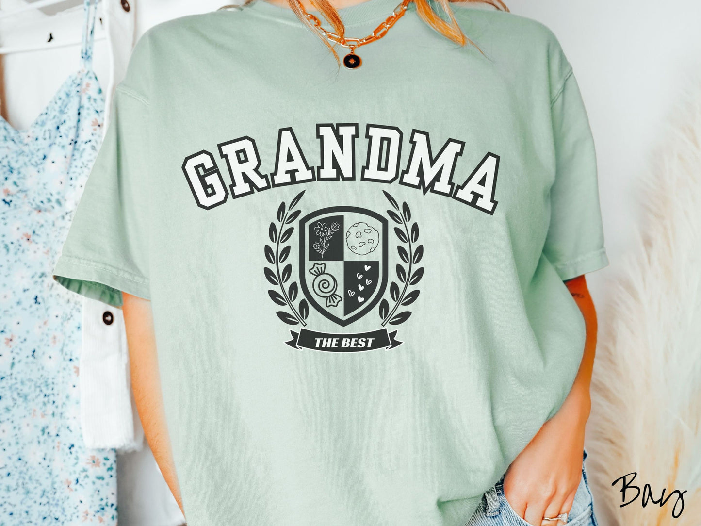 A woman wearing a cute bay colored shirt with text saying Grandma across the front with a varsity logo underneath with the words The Best centered underneath the logo.