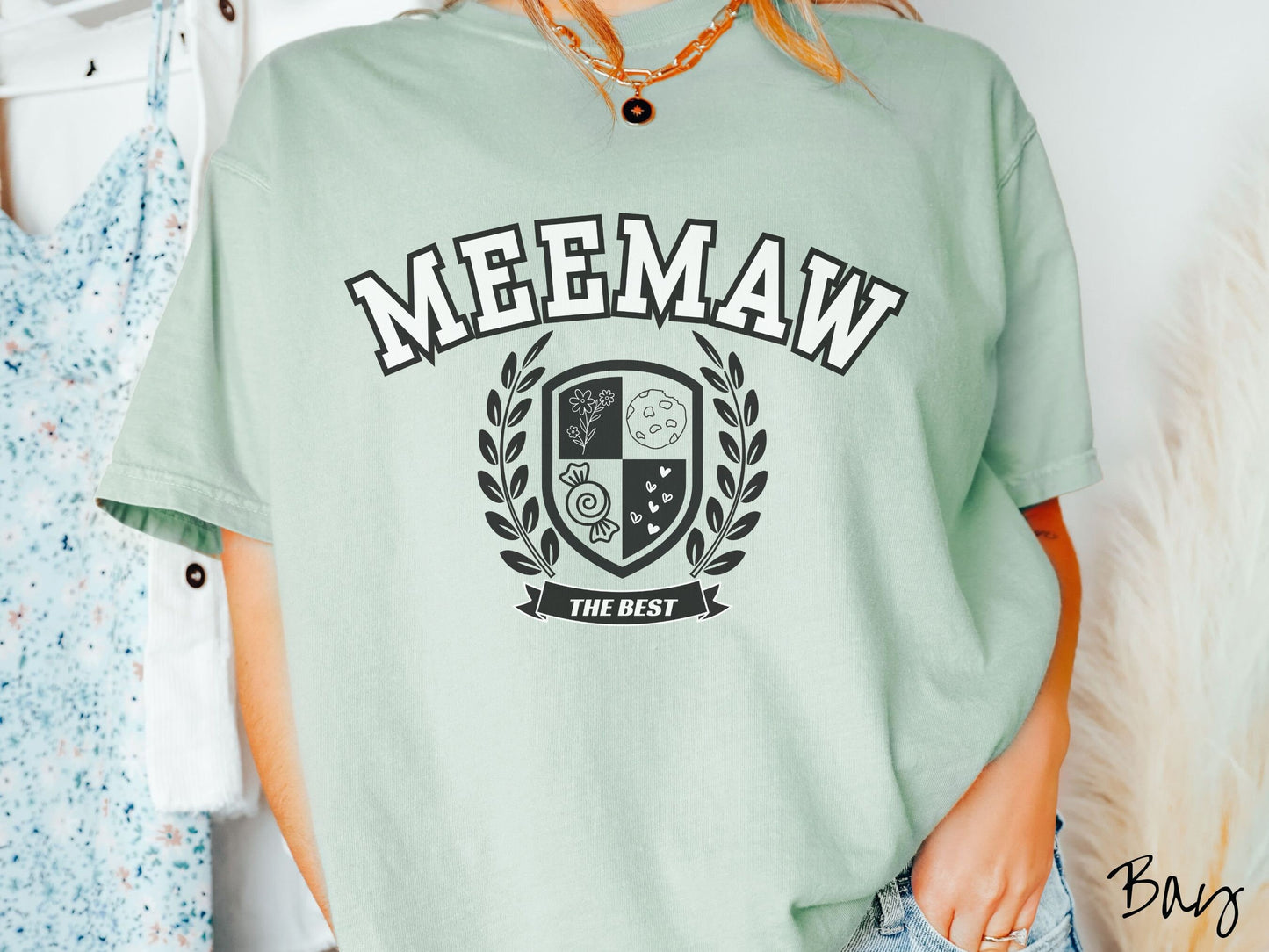 A woman wearing a cute bay colored shirt with text saying Meemaw across the front with a varsity logo underneath with the words The Best centered underneath the logo.