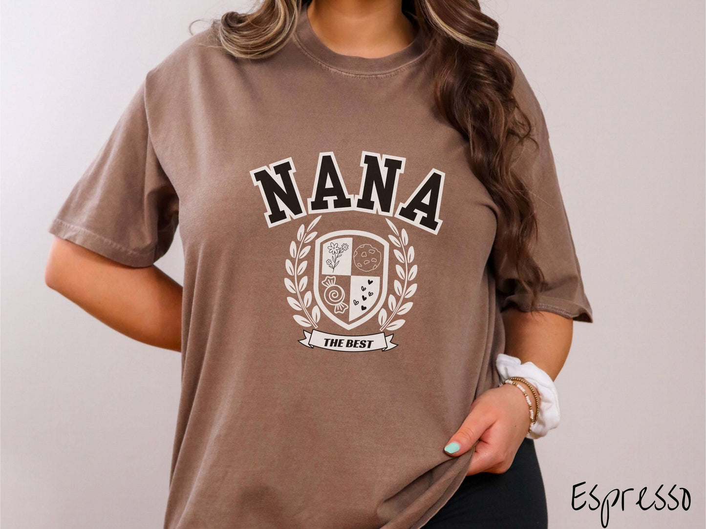 A woman wearing a cute espresso colored shirt with text saying Nana across the front with a varsity logo underneath with the words The Best centered underneath the logo.
