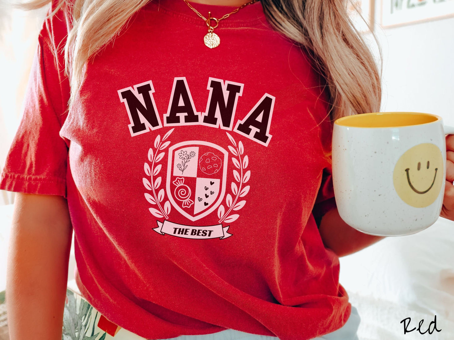 A woman wearing a cute red colored shirt with text saying Nana across the front with a varsity logo underneath with the words The Best centered underneath the logo.