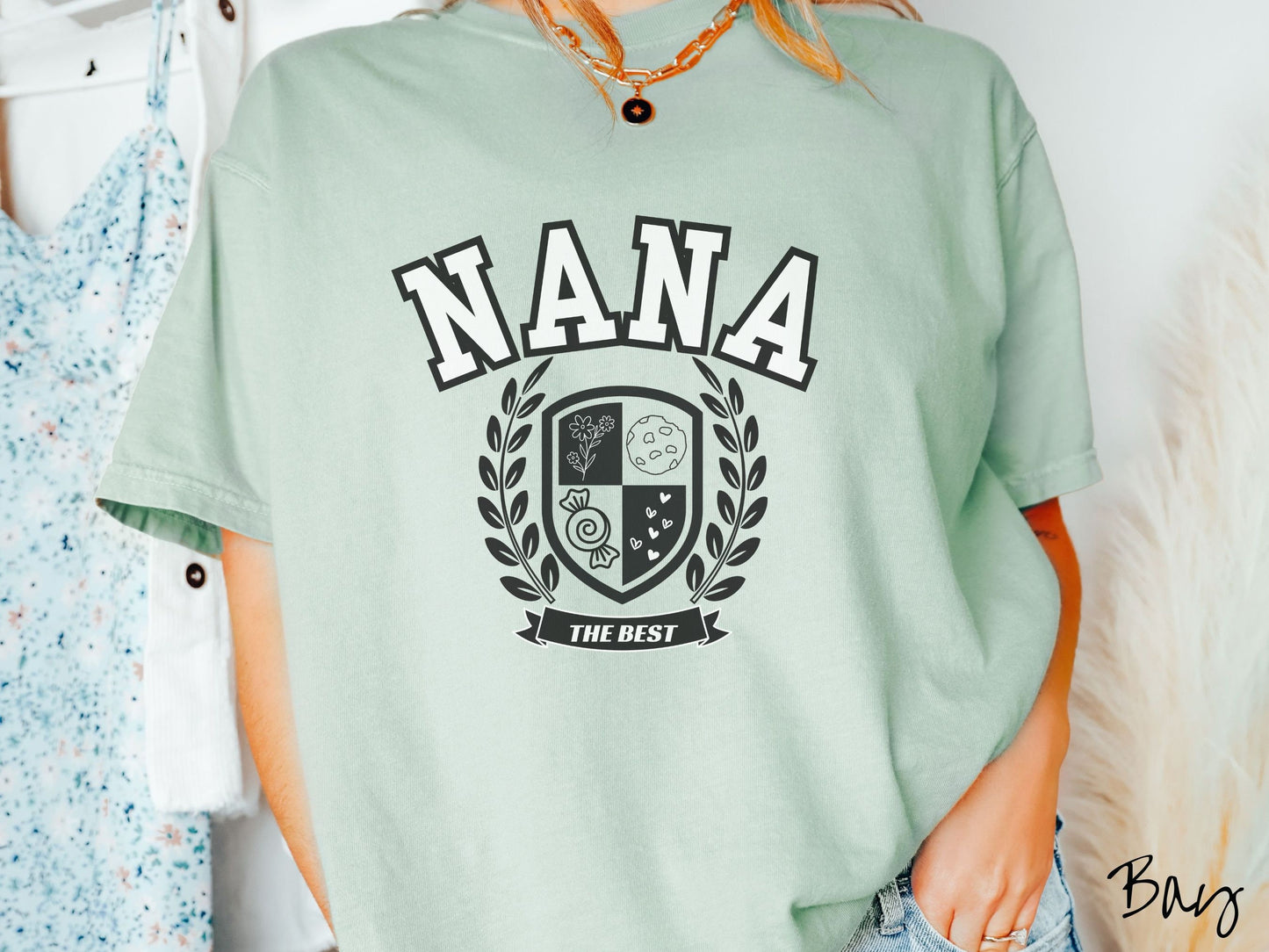 A woman wearing a cute bay colored shirt with text saying Nana across the front with a varsity logo underneath with the words The Best centered underneath the logo.