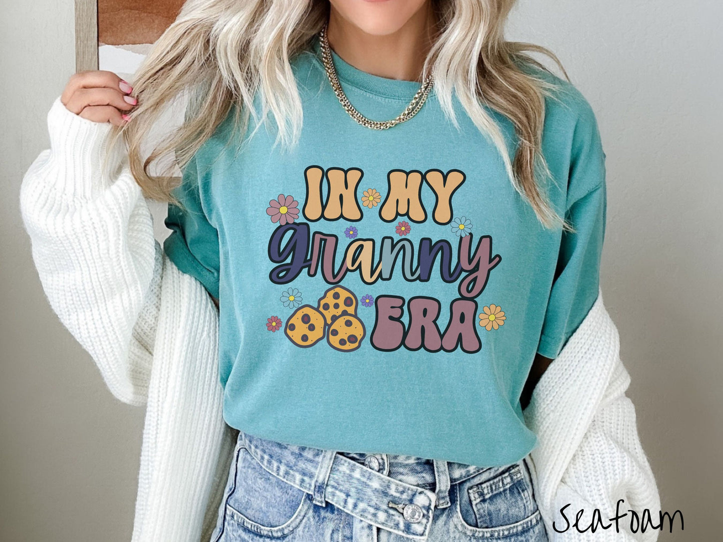 A woman wearing a cute seafoam colored sweatshirt with text on the front saying In My Granny Era, with flowers and chocolate chip cookies mixed within the text.