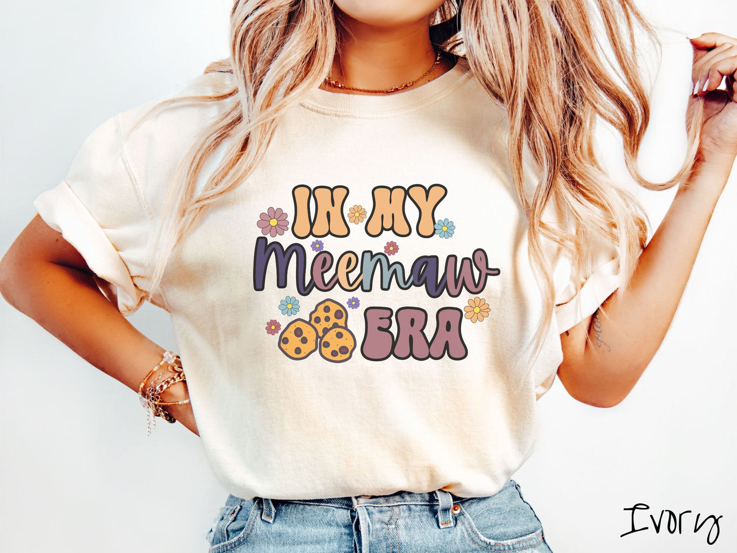 A woman wearing a cute ivory colored sweatshirt with text on the front saying In My Meemaw Era, with flowers and chocolate chip cookies mixed within the text.