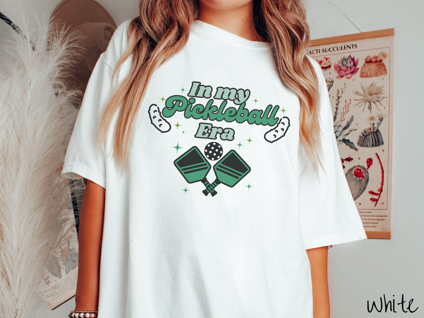 A woman wearing a cute white colored shirt with the text In My Pickleball Era across the top, and below are 2 pickleball paddles and a ball. There are pickles at the bottom left and right corners of the text.