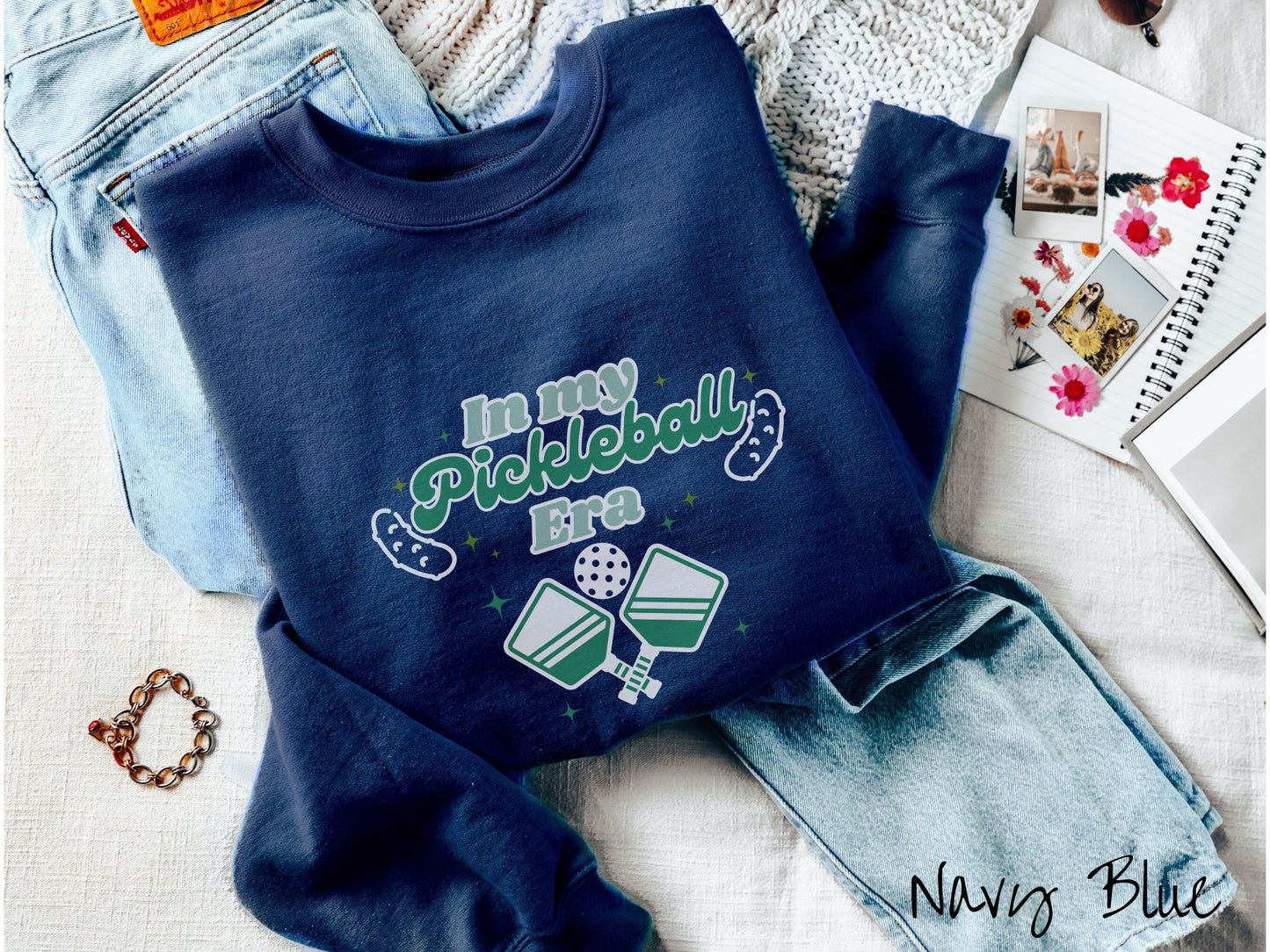 A woman wearing a cute navy blue colored sweatshirt with the text In My Pickleball Era across the top, and below are 2 pickleball paddles and a ball. There are pickles at the bottom left and right corners of the text.