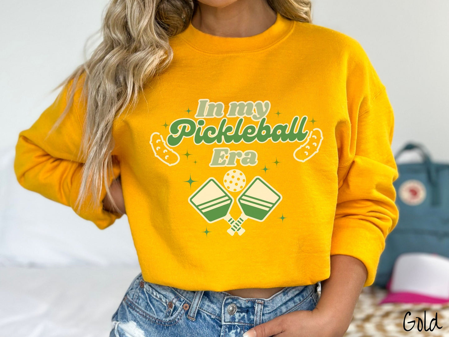 A woman wearing a cute gold colored sweatshirt with the text In My Pickleball Era across the top, and below are 2 pickleball paddles and a ball. There are pickles at the bottom left and right corners of the text.