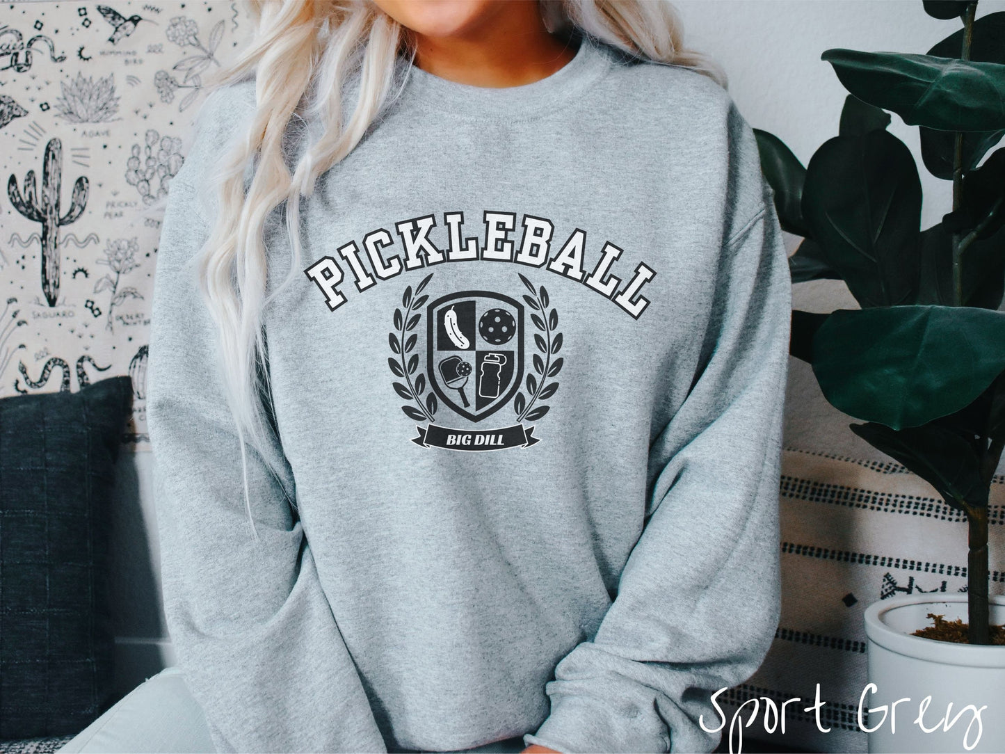 A woman wearing a cute sport grey colored sweatshirt with the text Pickleball across the top, a varsity logo centered underneath, and the words Big Dill underneath the logo.