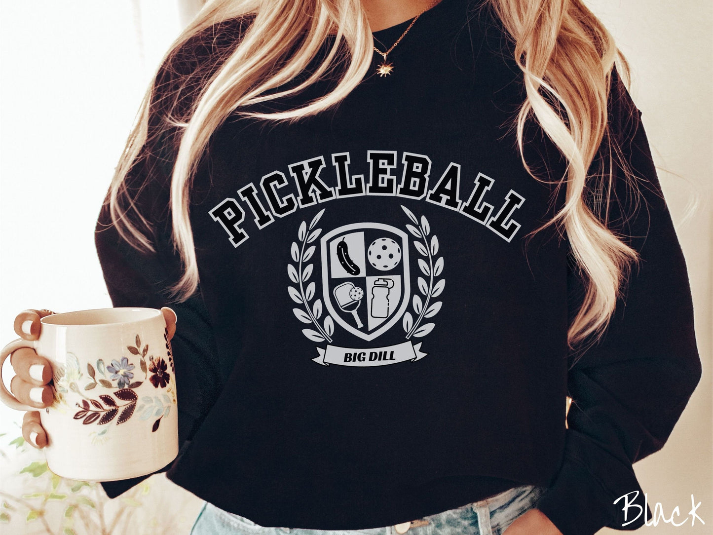 A woman wearing a cute black colored sweatshirt with the text Pickleball across the top, a varsity logo centered underneath, and the words Big Dill underneath the logo.