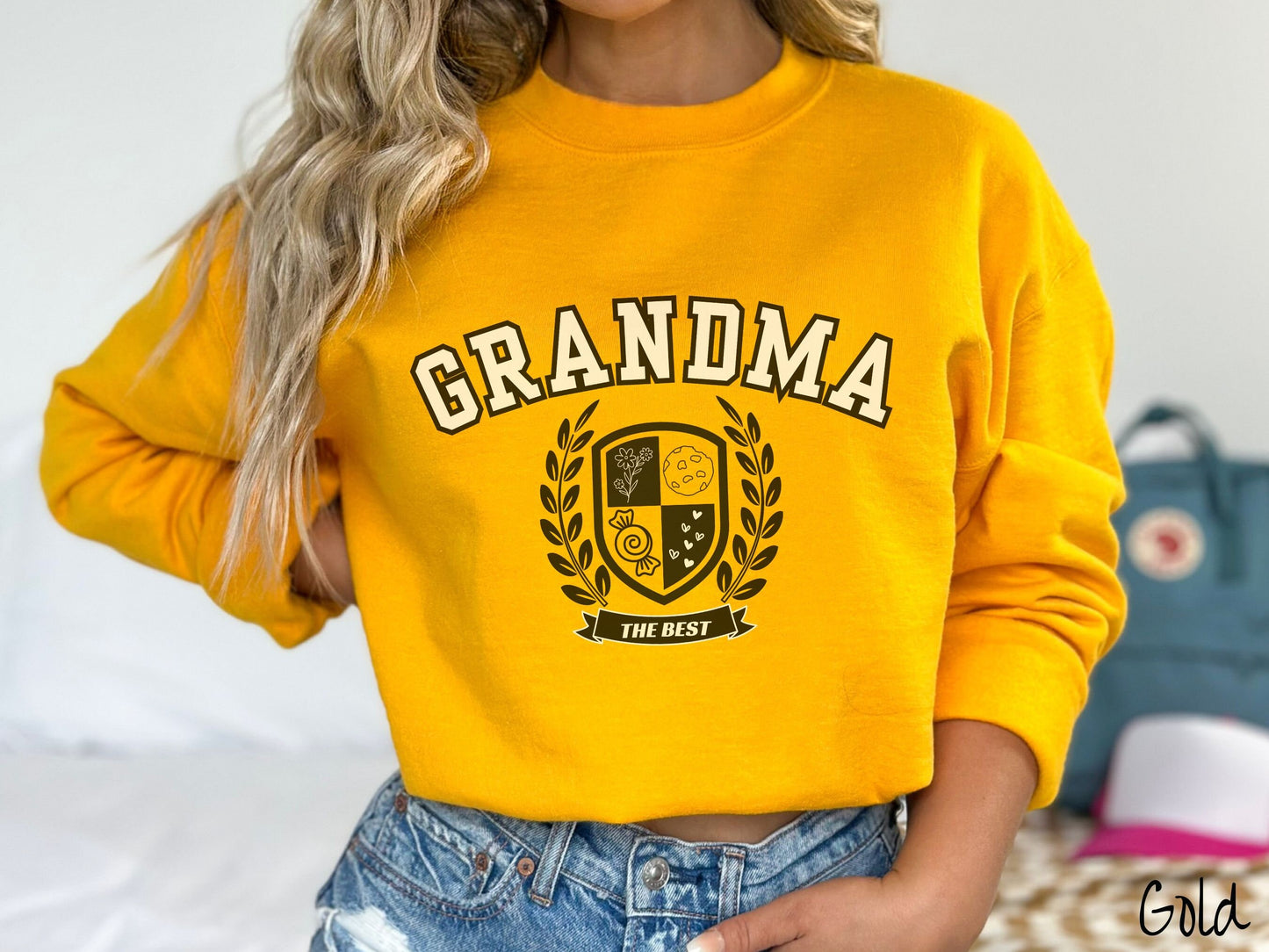A woman wearing a cute gold colored sweatshirt with text saying Grammy across the front with a varsity logo underneath with the words The Best centered underneath the logo.