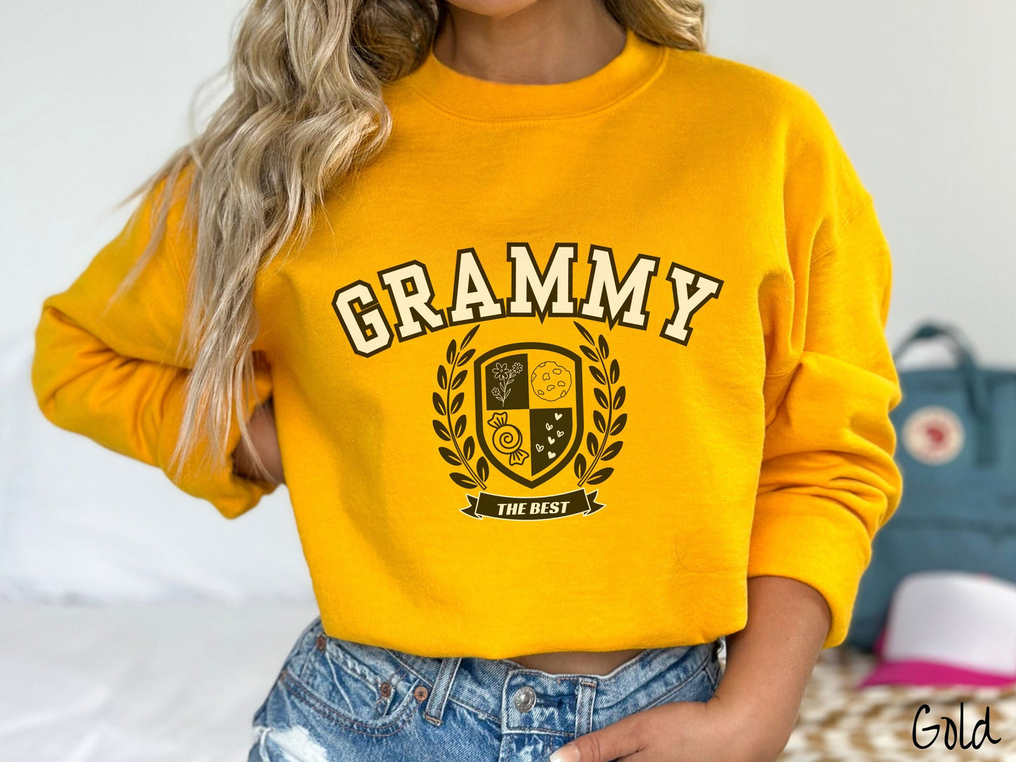 A woman wearing a cute gold colored sweatshirt with text saying Grammy across the front with a varsity logo underneath with the words The Best centered underneath the logo.