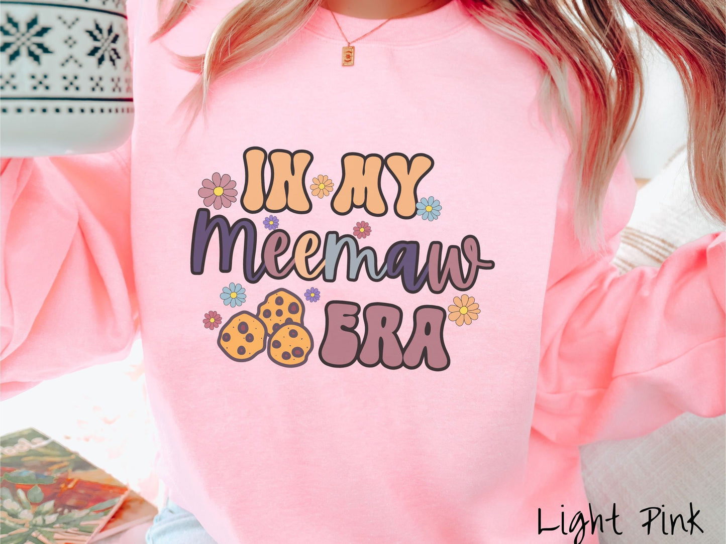 A woman wearing a cute light pink colored sweatshirt with text on the front saying In My Grandmother Era, with flowers and chocolate chip cookies mixed within the text.