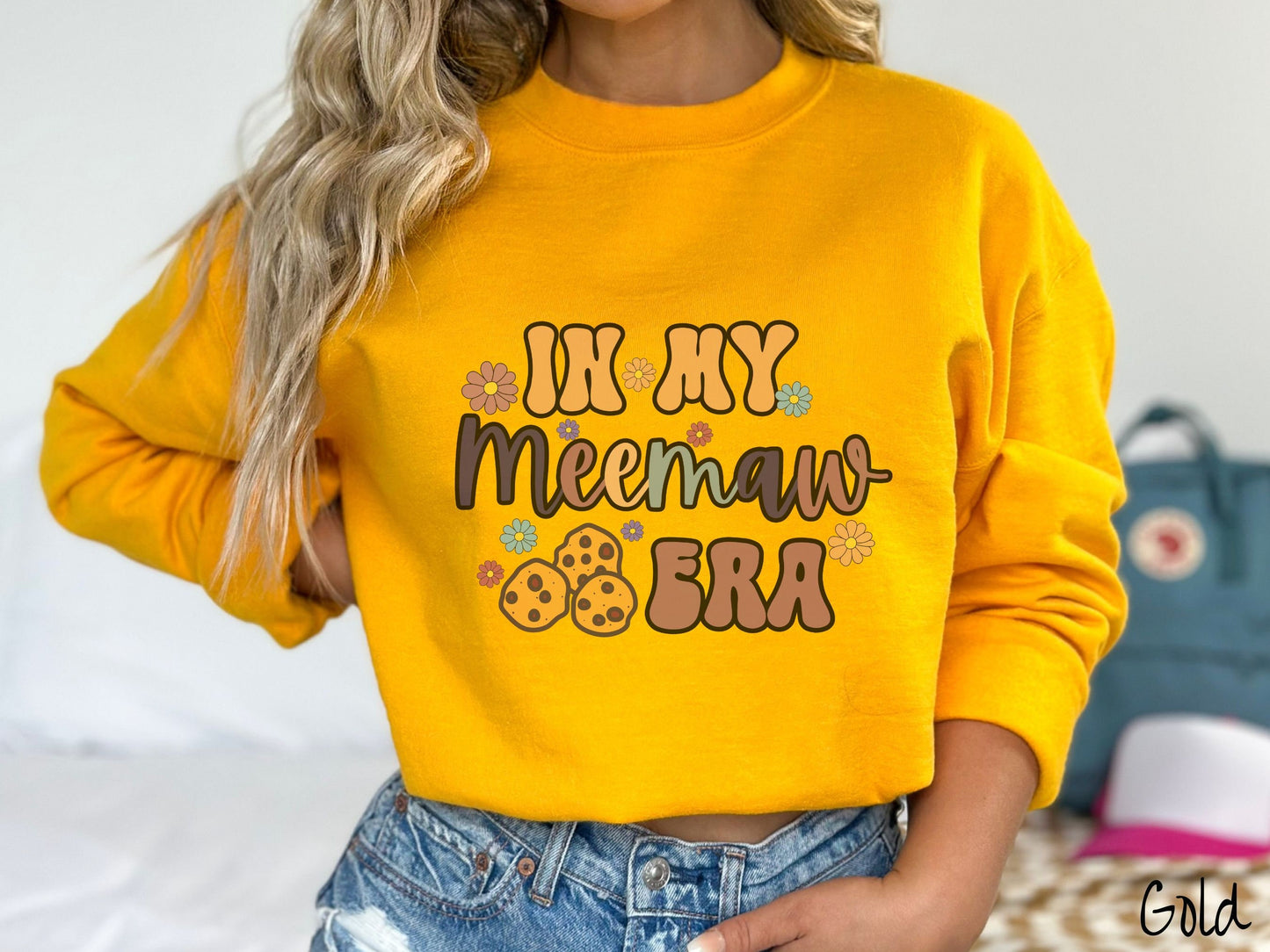 A woman wearing a cute gold colored sweatshirt with text on the front saying In My Grandmother Era, with flowers and chocolate chip cookies mixed within the text.