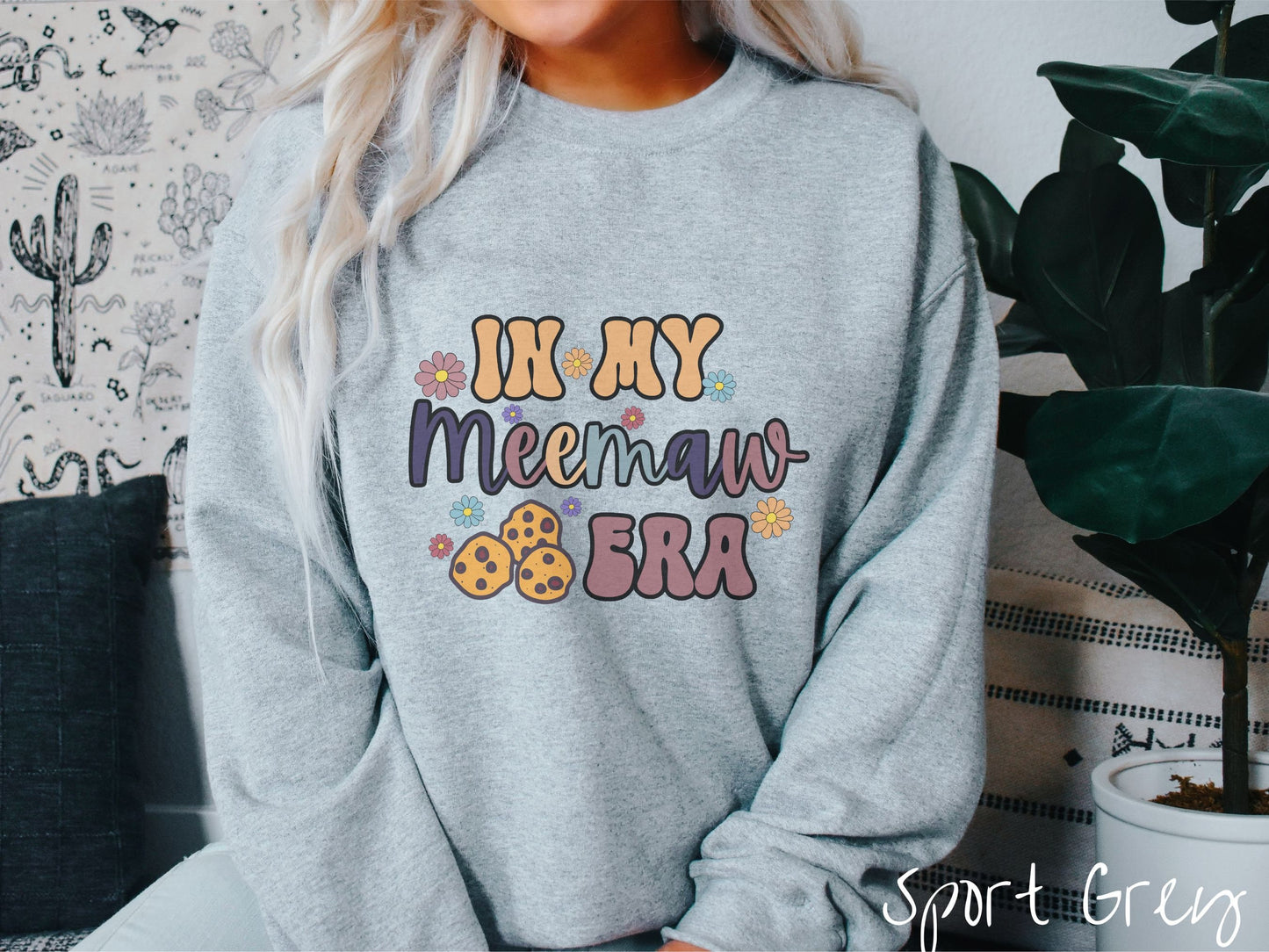 A woman wearing a cute sport grey colored sweatshirt with text on the front saying In My Grandmother Era, with flowers and chocolate chip cookies mixed within the text.