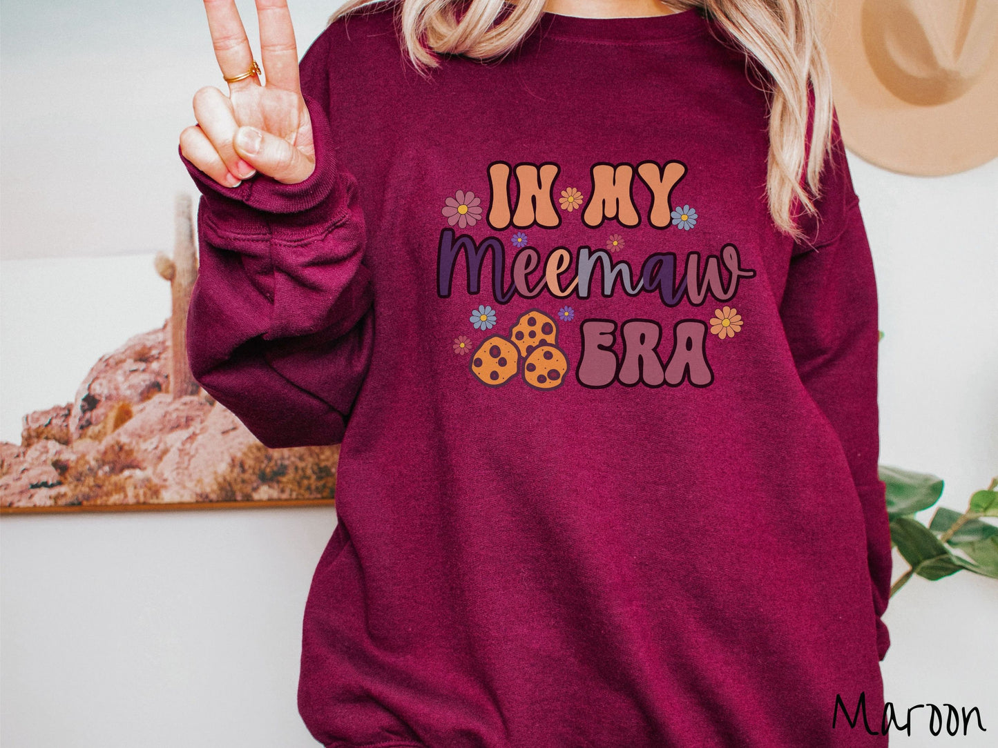 A woman wearing a cute maroon colored sweatshirt with text on the front saying In My Grandmother Era, with flowers and chocolate chip cookies mixed within the text.