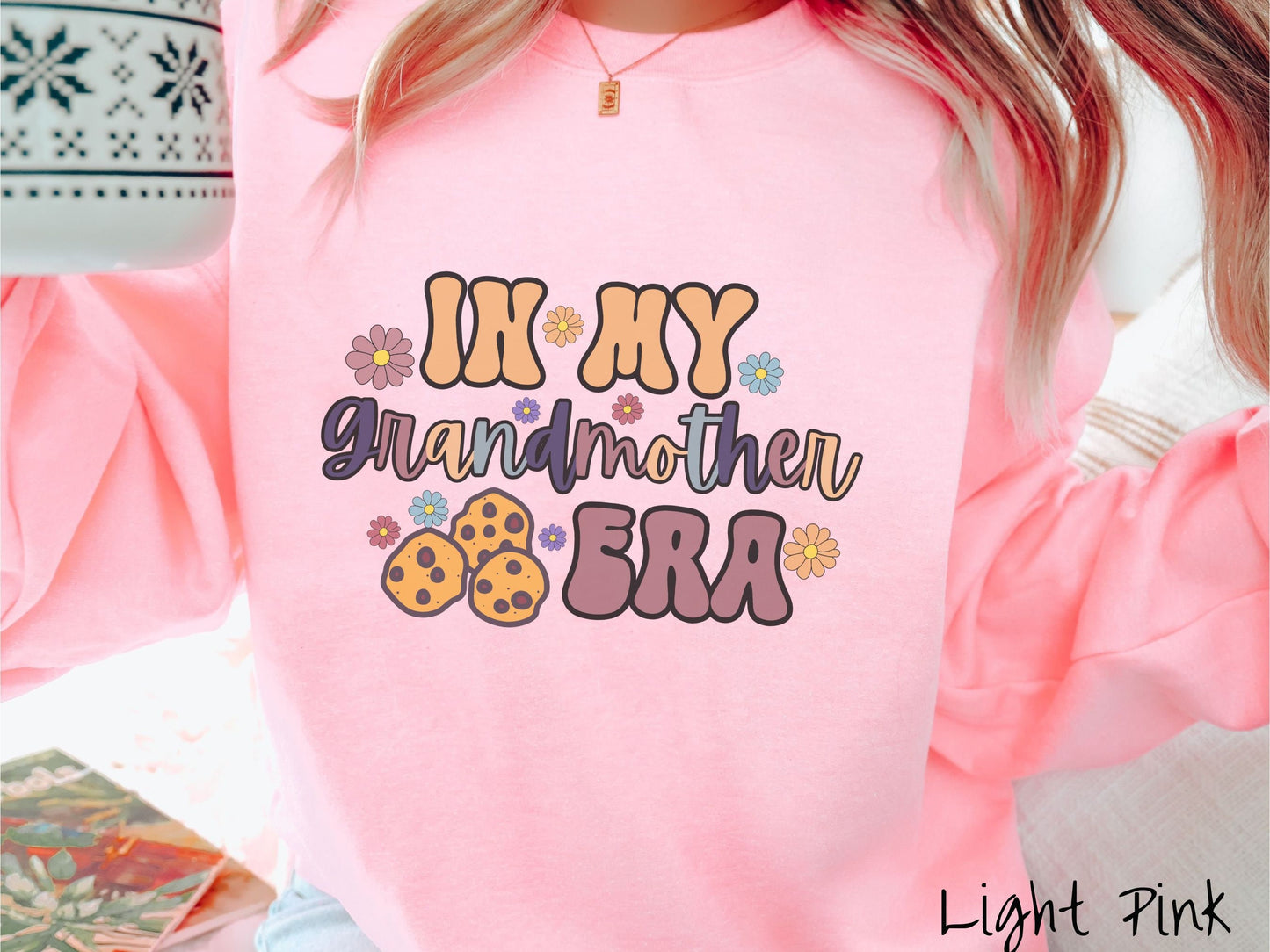 A woman wearing a cute light pink colored sweatshirt with text on the front saying In My Grandmother Era, with flowers and chocolate chip cookies mixed within the text.