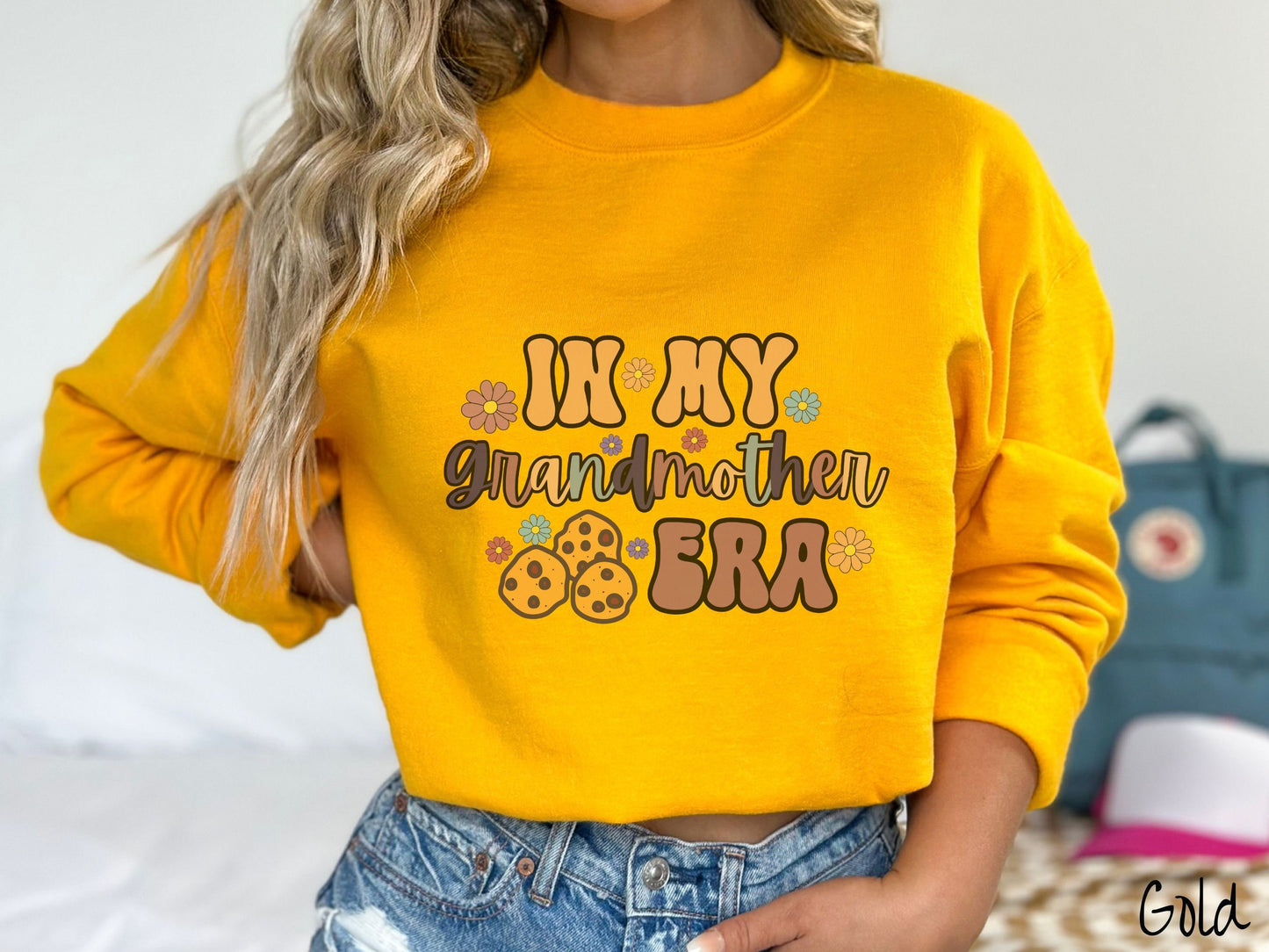 A woman wearing a cute gold colored sweatshirt with text on the front saying In My Grandmother Era, with flowers and chocolate chip cookies mixed within the text.